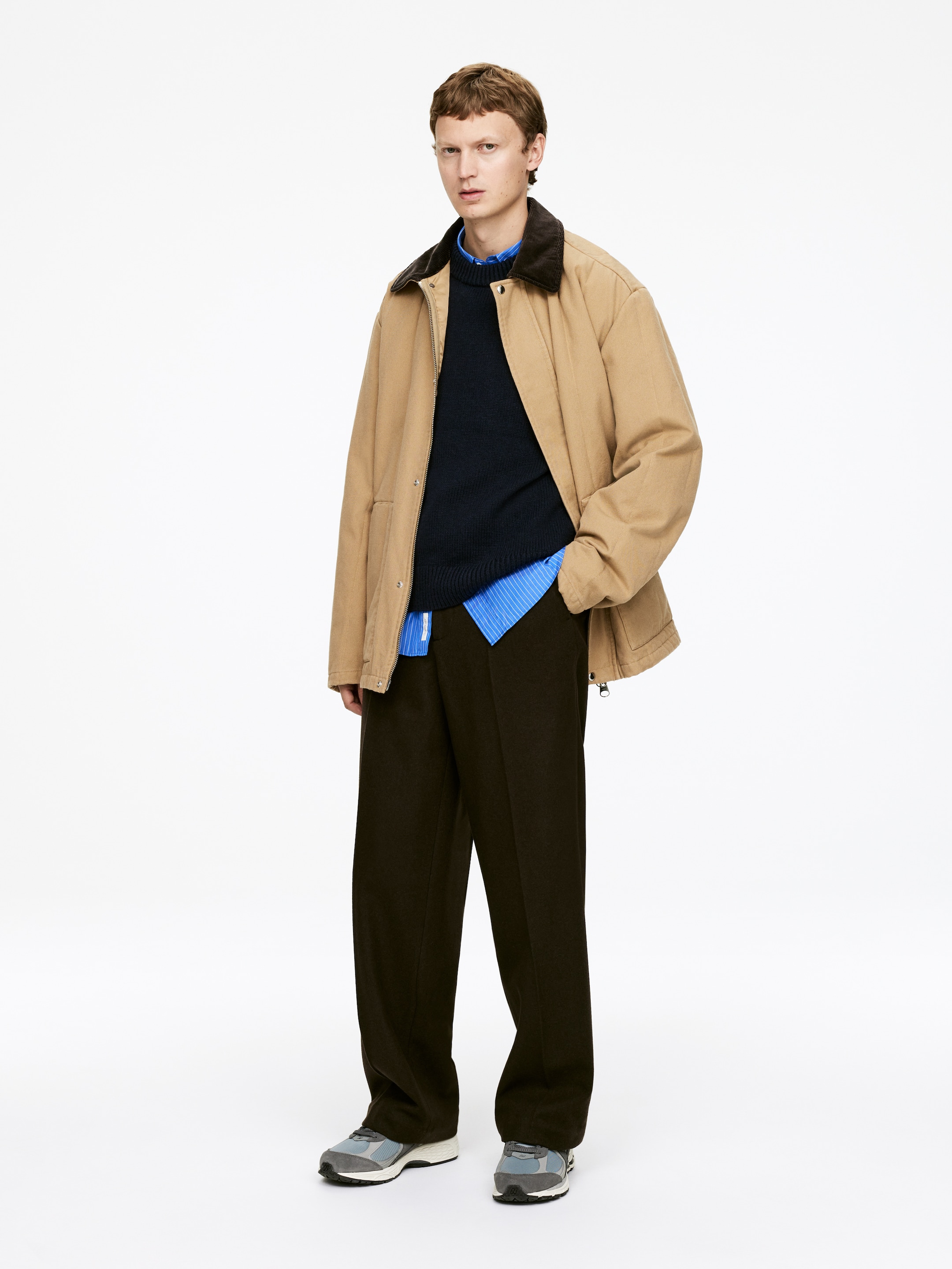 View larger image: A person is standing facing forward, modeling an outfit featuring a tan workwear-style jacket with a dark brown corduroy collar, layered over a navy crew-neck sweater and a light blue striped collared shirt. Dark brown wide-leg trousers are completing the look, paired with grey and light blue athletic sneakers.