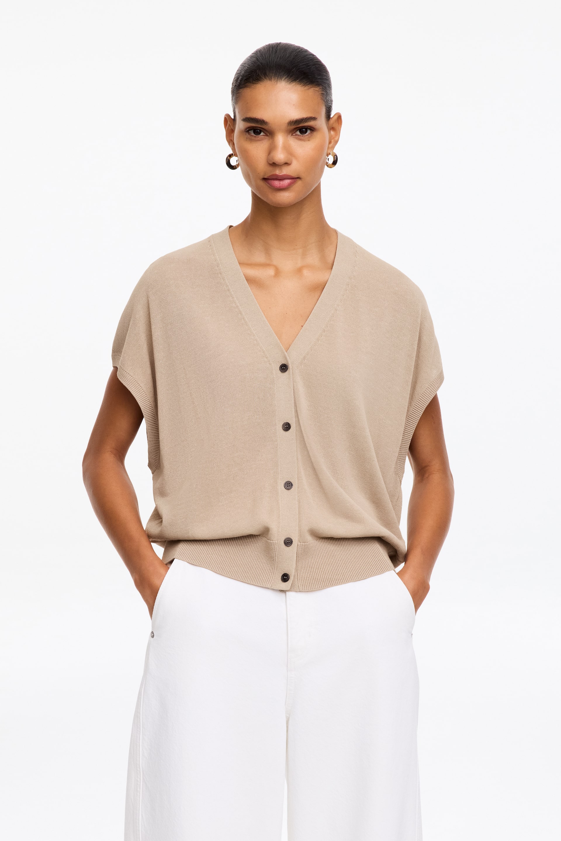 Grotere afbeelding bekijken: Een beige gebreid vest met korte mouwen wordt frontaal gepresenteerd, met een V-hals, een vijfknoopssluiting aan de voorkant met donkere knopen, korte kapmouwtjes en een geribde zoom.