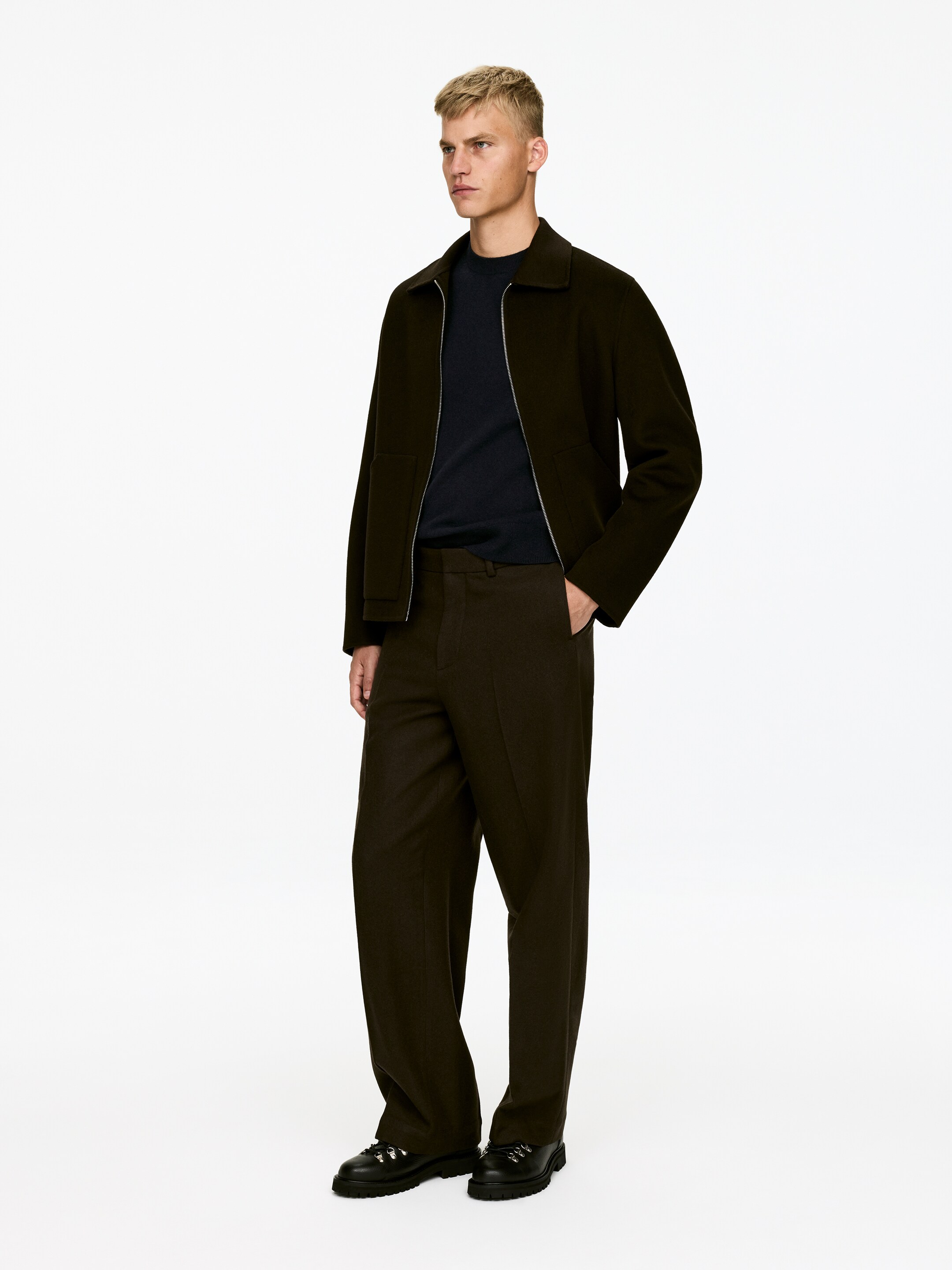 Agrandir l'image: Une veste marron foncé à col, avec une fermeture éclair apparente de couleur argentée et deux poches avant horizontales, est portée ouverte sur un pull à col rond bleu foncé. Un pantalon large marron foncé assorti complète l'ensemble, qui est également composé de chaussures noires à lacets avec semelles épaisses, et qui est orienté aux trois quarts vers la gauche.