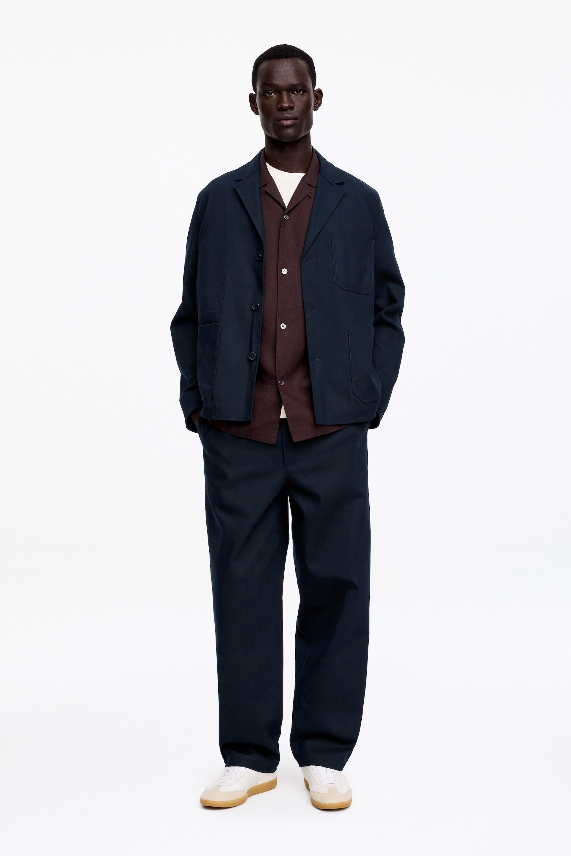 Se større billede: Et komplet outfit præsenteres, bestående af en mørkeblå jakke med notch-revers, tre forknapper og to påsyede lommer. Jakken bæres over en åben brun skjorte med camp-krave, som afslører en hvid crew neck t-shirt underneden. Matchende mørkeblå, løstsiddende bukser fuldender ensemblet, og lyse sneakers med gummisåler sætter prikken over i'et.