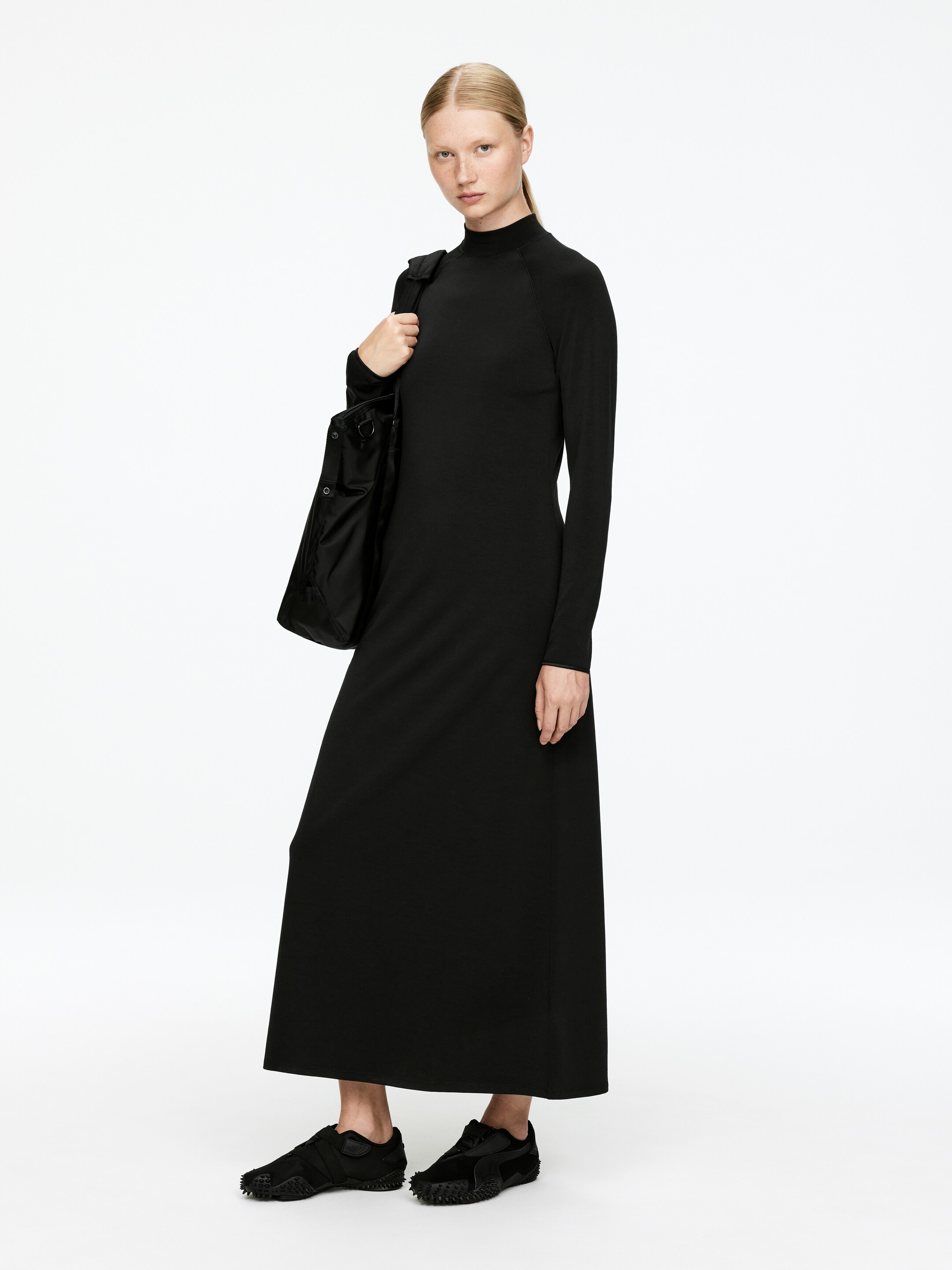 Vezi imaginea mai mare: O rochie neagră, lungă până la podea, cu mâneci lungi, guler rulat și siluetă subțire este purtată de model. Modelul poartă o geantă de umăr neagră și pantofi negri.