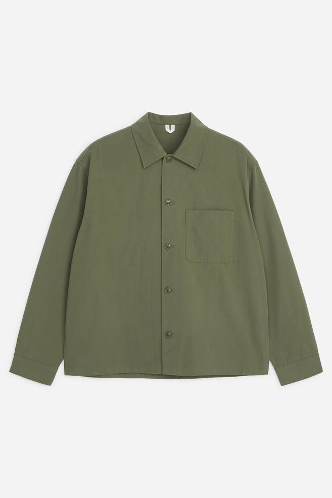 An olive green cotton overshirt is shown from the front, featuring a regular collar, a five-button front fastening, and a single patch pocket on the chest. The long-sleeved garment is presented in a relaxed, regular fit with structured cuffs. - 1