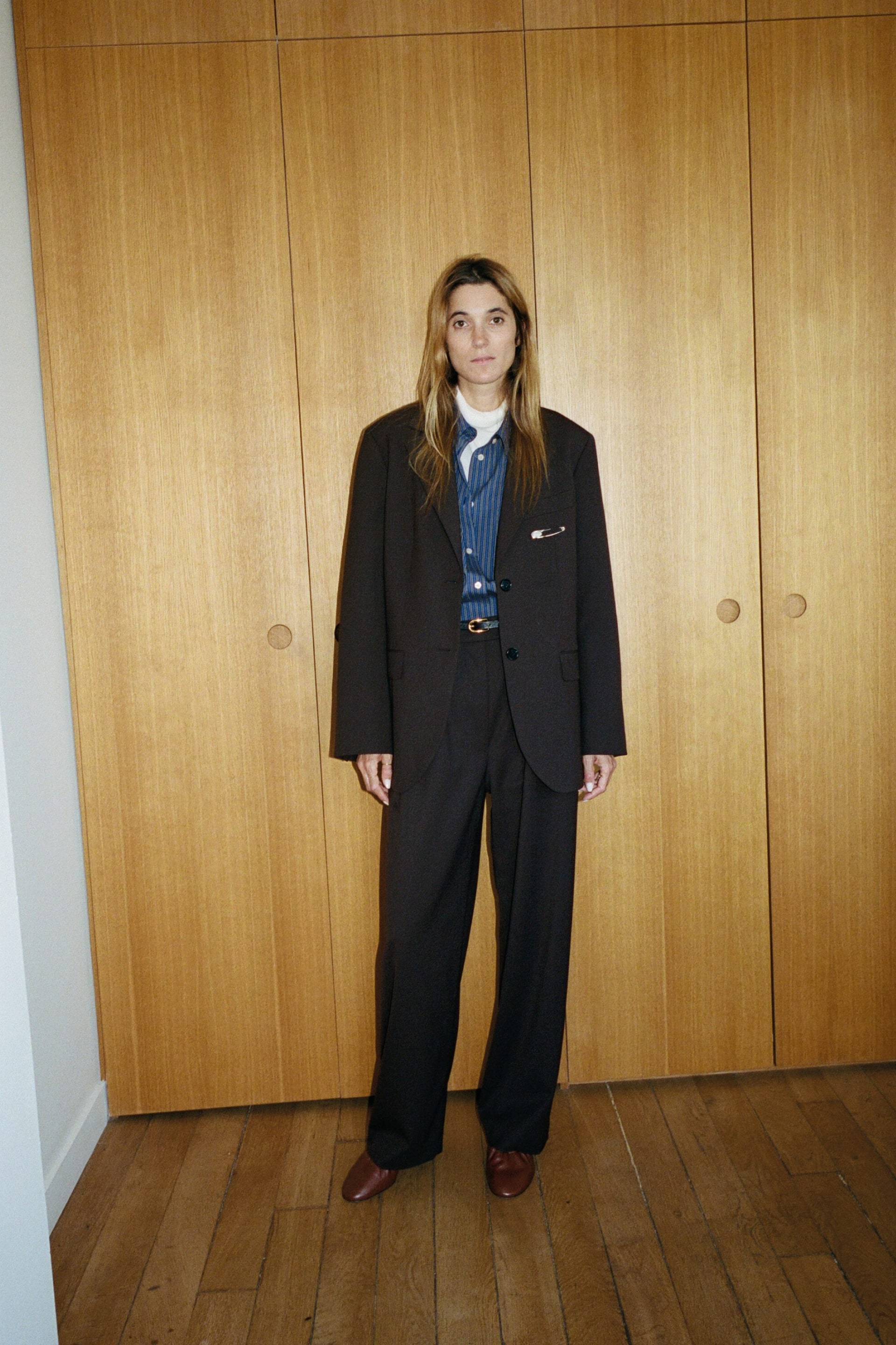Visualizza l'immagine ingrandita: Un modello è in piedi rivolto frontalmente, indossando un blazer sartoriale marrone scuro e pantaloni a gamba larga coordinati, abbinati a una camicia a righe blu sovrapposta a un maglione dolcevita bianco. Una spilla brooch placcata in argento dettaglia il taschino del blazer, mentre una sottile cintura in pelle con fibbia dorata segna il punto vita e delle décolleté in pelle marrone completano l’outfit.