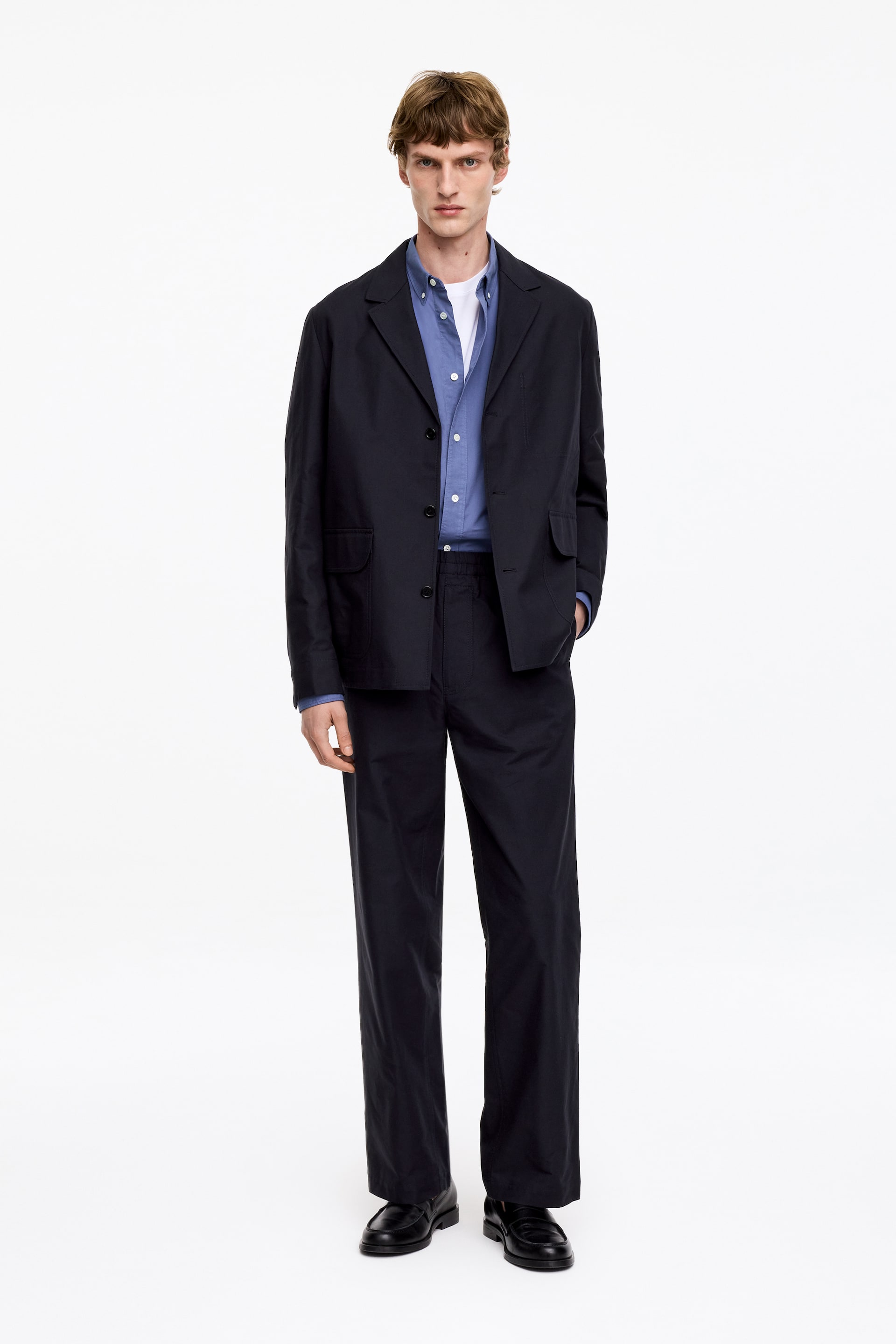 Agrandir l'image: Le modèle porte un blazer bleu foncé à simple boutonnage avec des revers crantés et deux poches plaquées, sur une chemise bleu clair et un t-shirt blanc. Il porte également un pantalon droit assorti bleu foncé avec des mocassins noirs, une main dans la poche du pantalon.