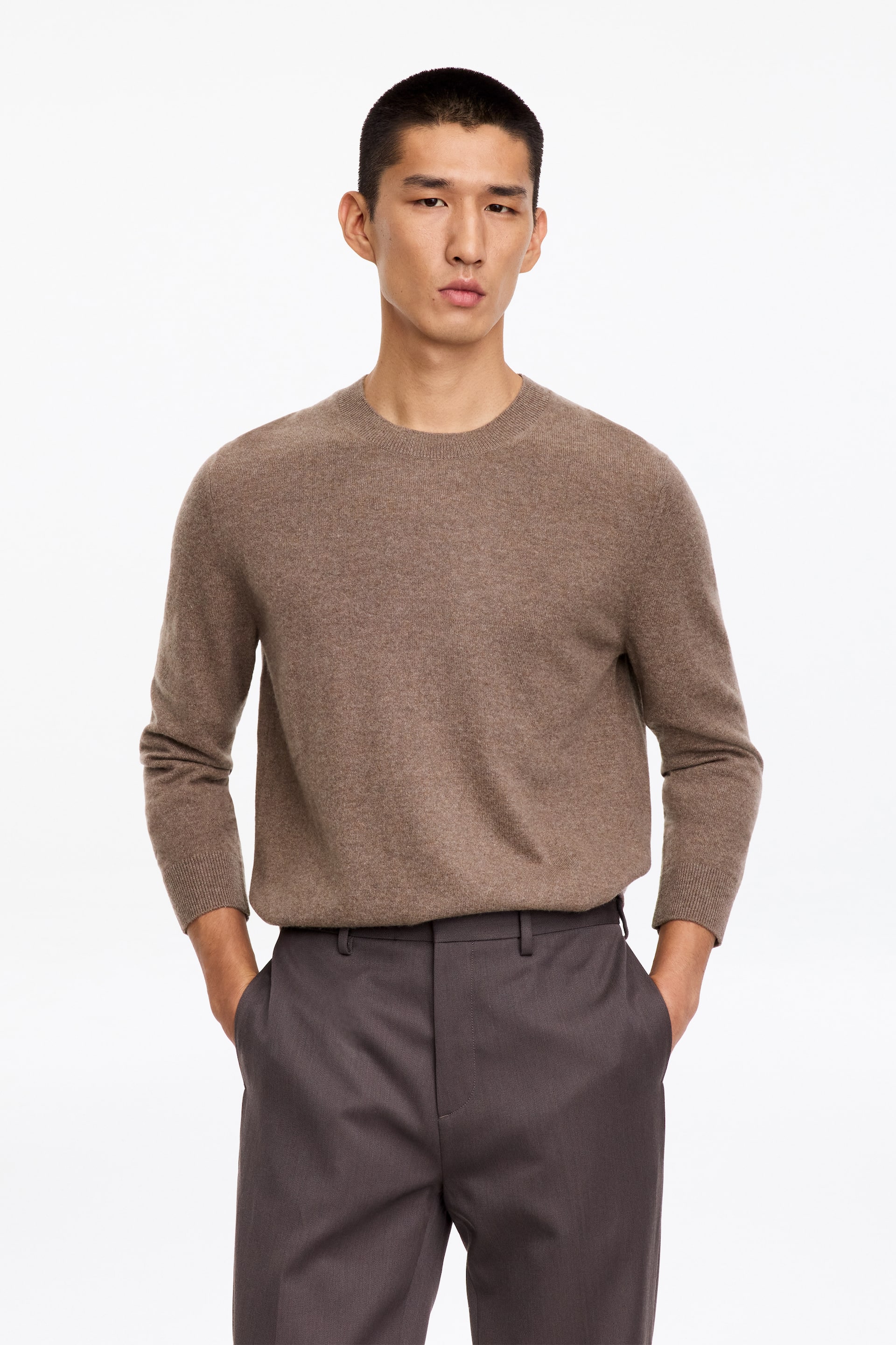 Visualizza immagine più grande: Il modello indossa un maglione marrone con girocollo, caratterizzato da collo e polsini a coste. Il maglione è infilato in un paio di pantaloni grigio scuro.