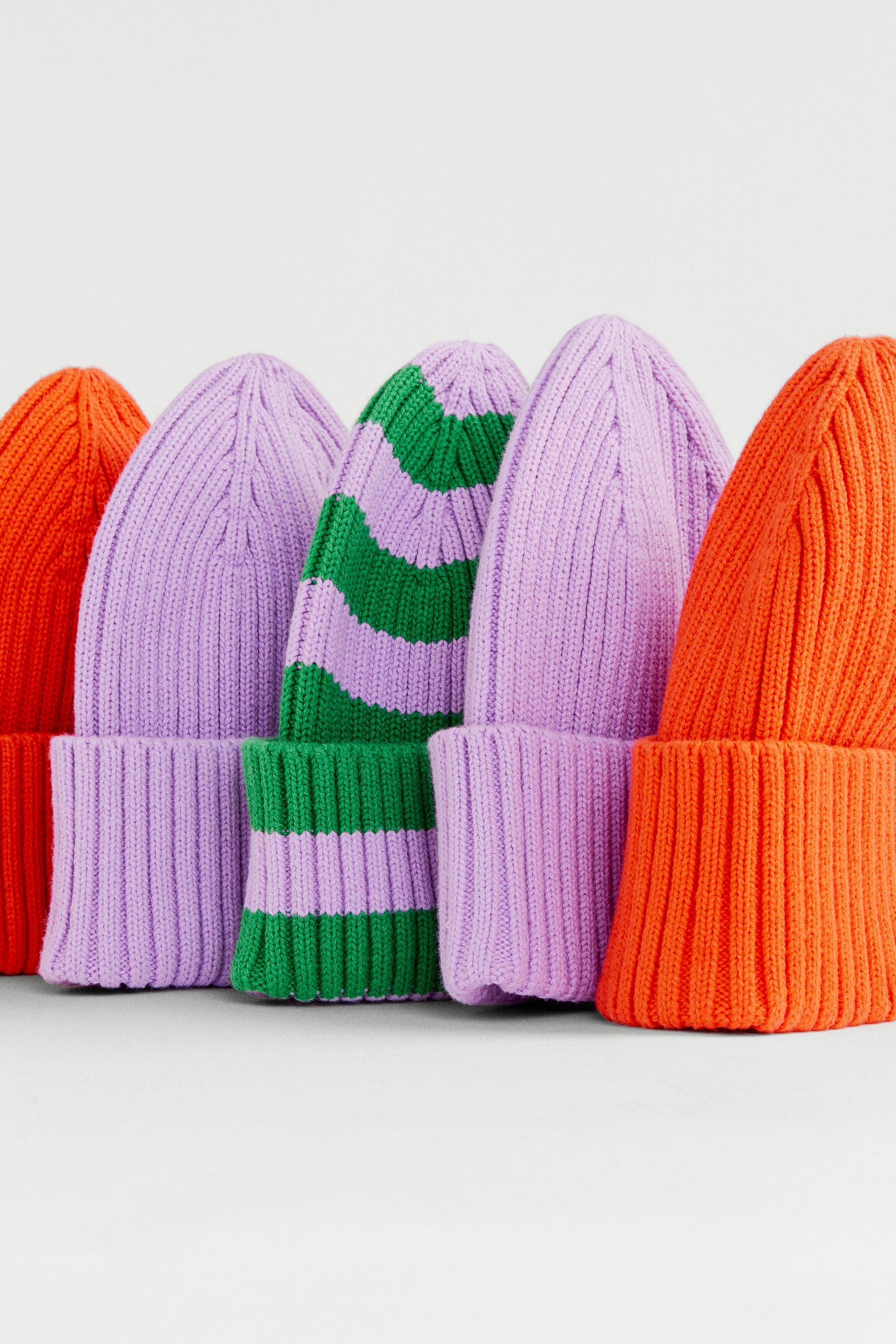 Grösseres Bild ansehen: Fünf Rippstrick-Beanies stehen aufrecht und zeigen ihre Vorderansicht. Sie präsentieren verschiedene Farben, darunter einfarbiges Orange, einfarbiges Lavendel sowie ein grün-lavendelfarbig gestreiftes Muster, jeweils mit umgeschlagenem Bund.