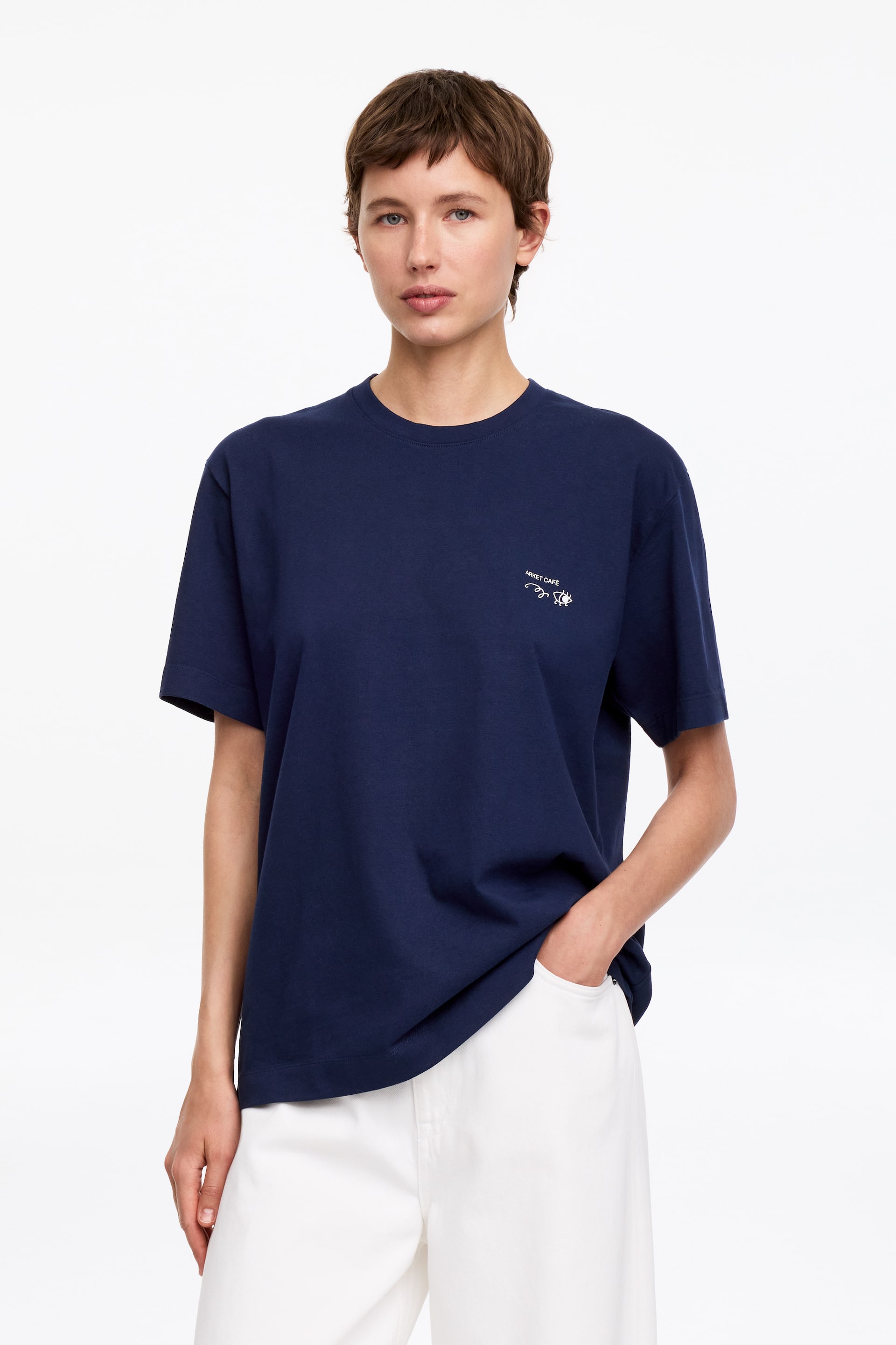 View larger image: A navy blue short-sleeved t-shirt featuring a crew neck and a small white embroidered "SUNSET CAFE" text with a sun graphic on the left chest. It displays a relaxed fit, with its hem partially tucked into white trousers on one side and hanging loose on the other.