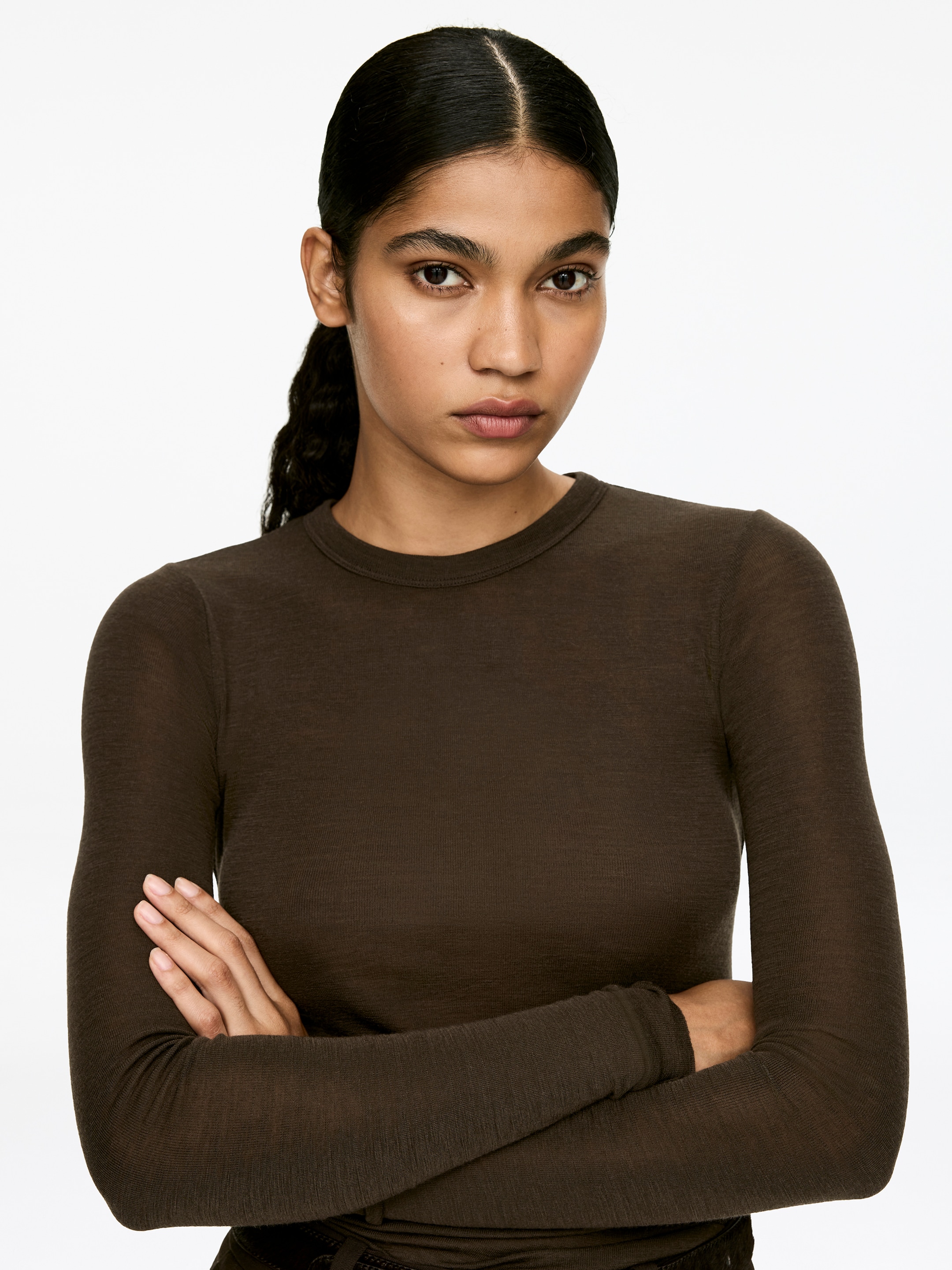 View larger image: A dark brown, tight-fitted, long-sleeved top, made of soft merino wool with a slightly transparent appearance, is being worn by the model. The model's arms are crossed, showcasing the garment's close fit.