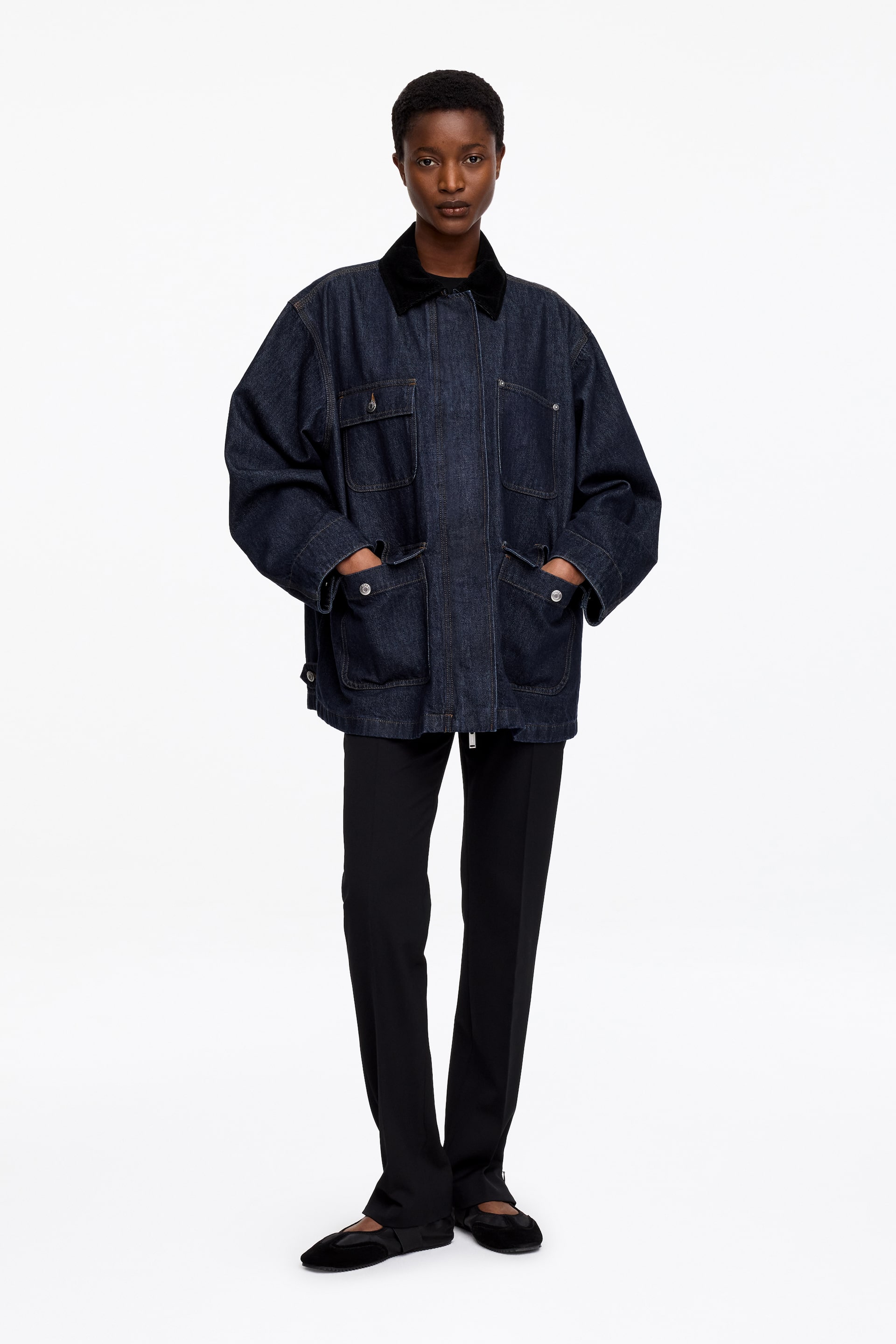 View larger image: The model is wearing an oversized denim jacket with a corduroy collar, with their hands resting in the lower patch pockets. The jacket boasts a zip closure with a placket, four front patch pockets with flaps, and button fastenings on three of them, along with side tabs featuring denim shank buttons.