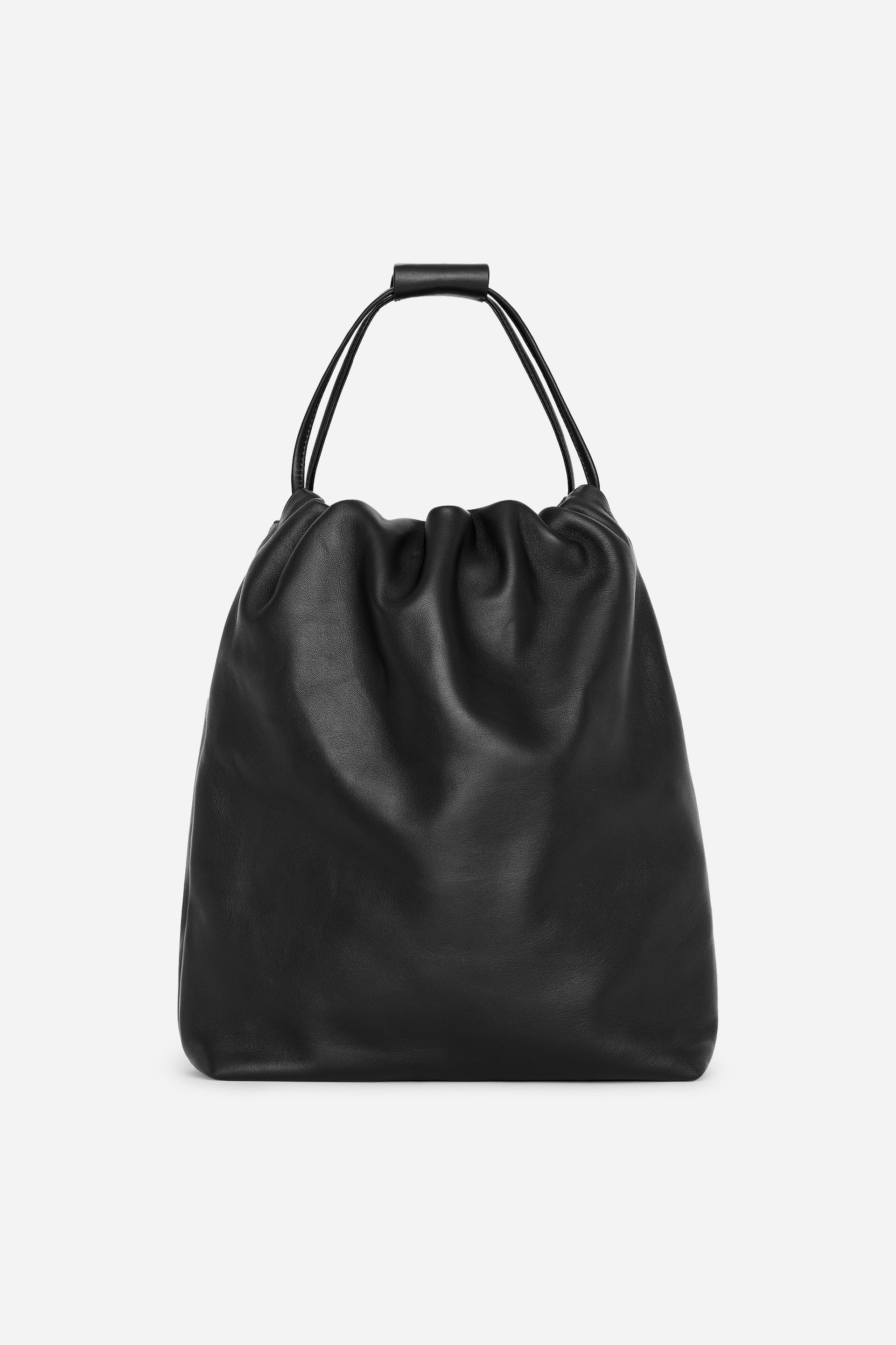 View larger image: A black leather shopping bag is shown from the front, featuring a slouchy silhouette with a gathered top opening. The slim drawstring-style shoulder straps extend upwards and are held together by a matching leather grip at the top.