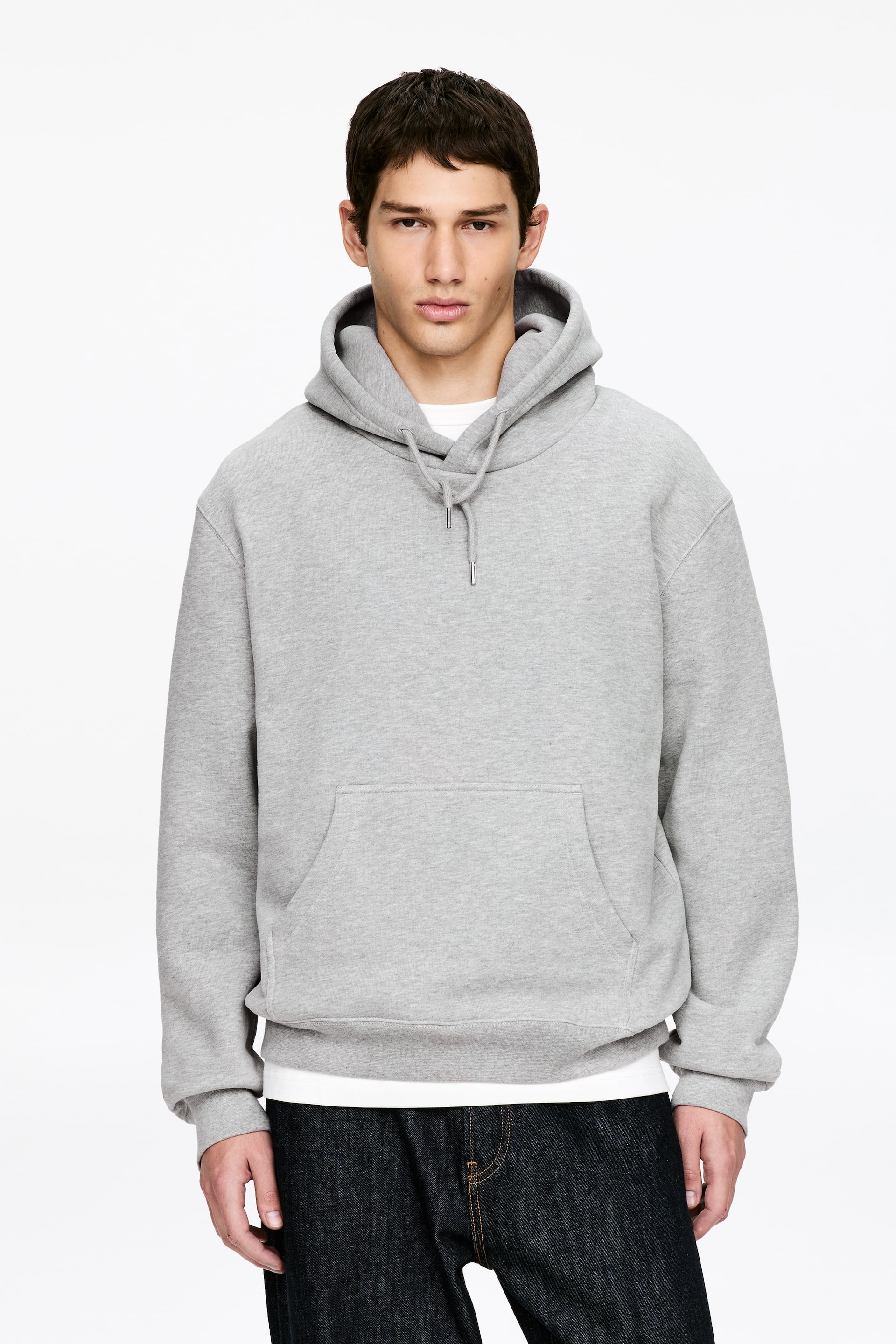 View larger image: A grey hoodie is facing forward, presenting a relaxed fit with a large hood, light drawstrings tipped with metal, and a front kangaroo pocket. It also features ribbed cuffs and a ribbed hem, with a white t-shirt layered underneath and dark jeans visible below.