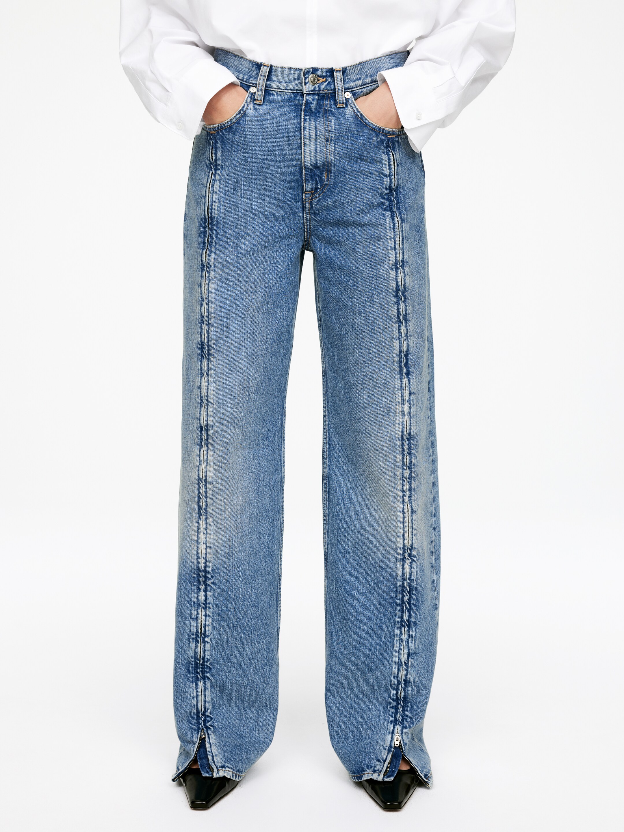 Visualizza immagine più grande: Un paio di jeans in denim blu con un lavaggio medio e un taglio a gamba larga viene presentato dalla parte anteriore, con una camicia bianca infilata nella cintura e le mani appoggiate nelle tasche anteriori. Cuciture verticali prominenti corrono lungo la parte anteriore di ciascuna gamba, e le cuciture interne delle caviglie mostrano cerniere aperte, rivelando scarpe nere.