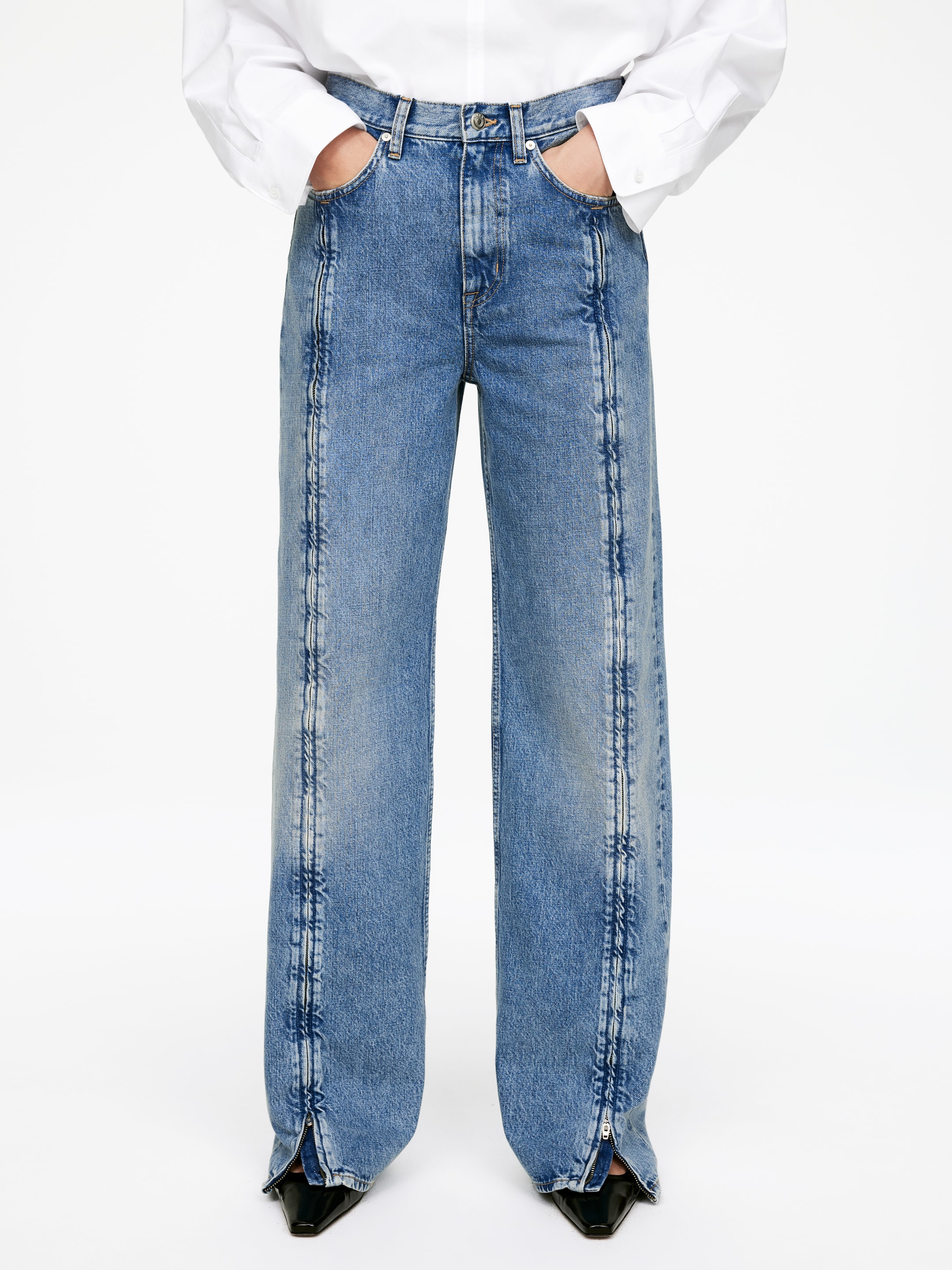 Visa större bild: Ett par blå denimjeans med en medelblå tvätt och vida ben presenteras framifrån, med en vit skjorta instoppad i midjan och händerna vilande i de främre fickorna. Framträdande vertikala sömmar löper längs framsidan av varje ben, och de inre ankelsömmarna visar öppna dragkedjor som avslöjar svarta skor.