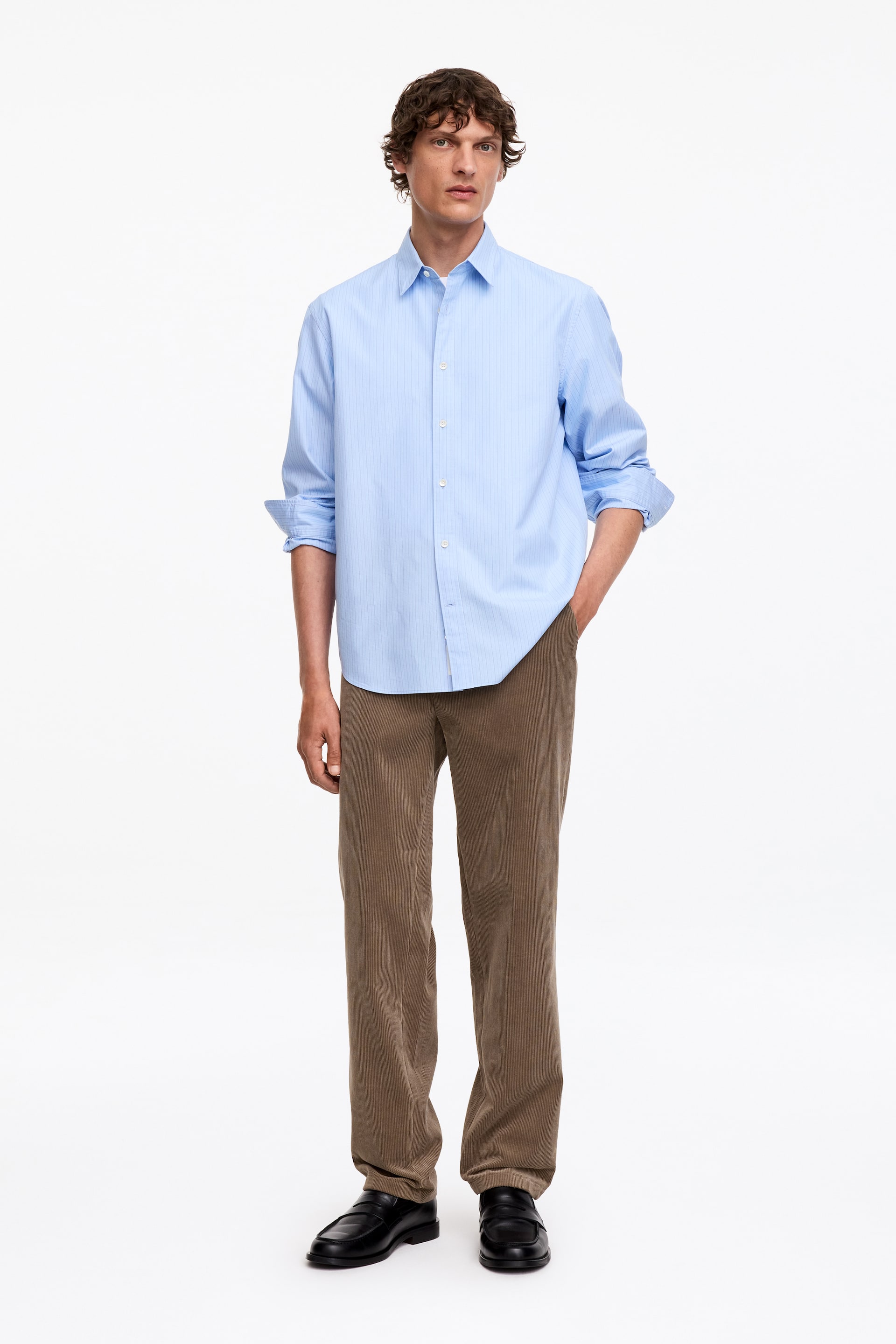 Ver imagen más grande: La modelo lleva puesta una camisa de botones de rayas azul claro. Presenta finas rayas verticales, un cuello clásico y mangas arremangadas hasta los antebrazos.