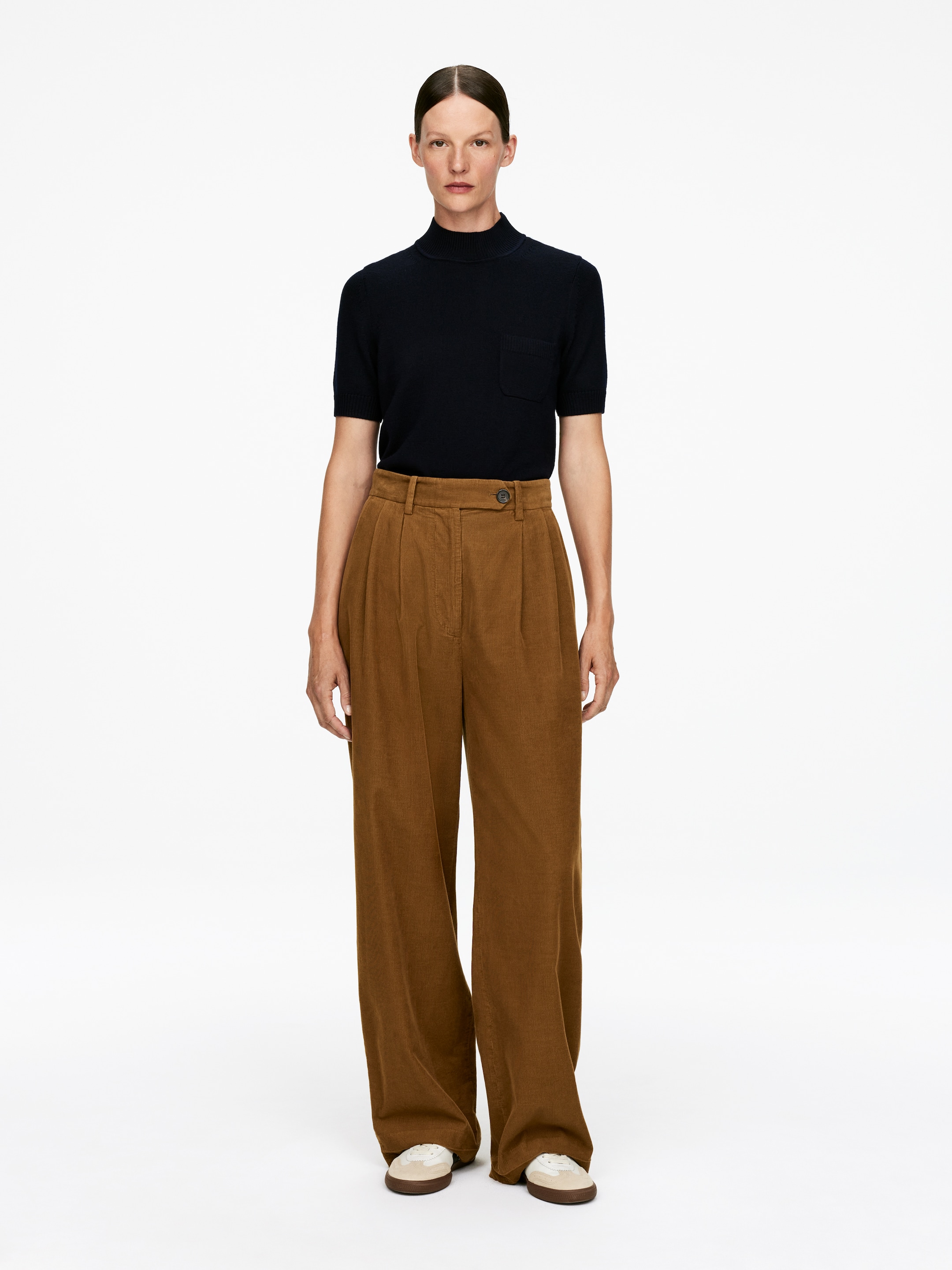 View larger image: A front-facing outfit is being presented, consisting of a dark navy short-sleeved mock-neck top with a chest patch pocket, which is being tucked into high-waisted, pleated brown wide-leg corduroy pants secured by a visible button closure at the waist. Light-colored sneakers with dark soles are completing the look.