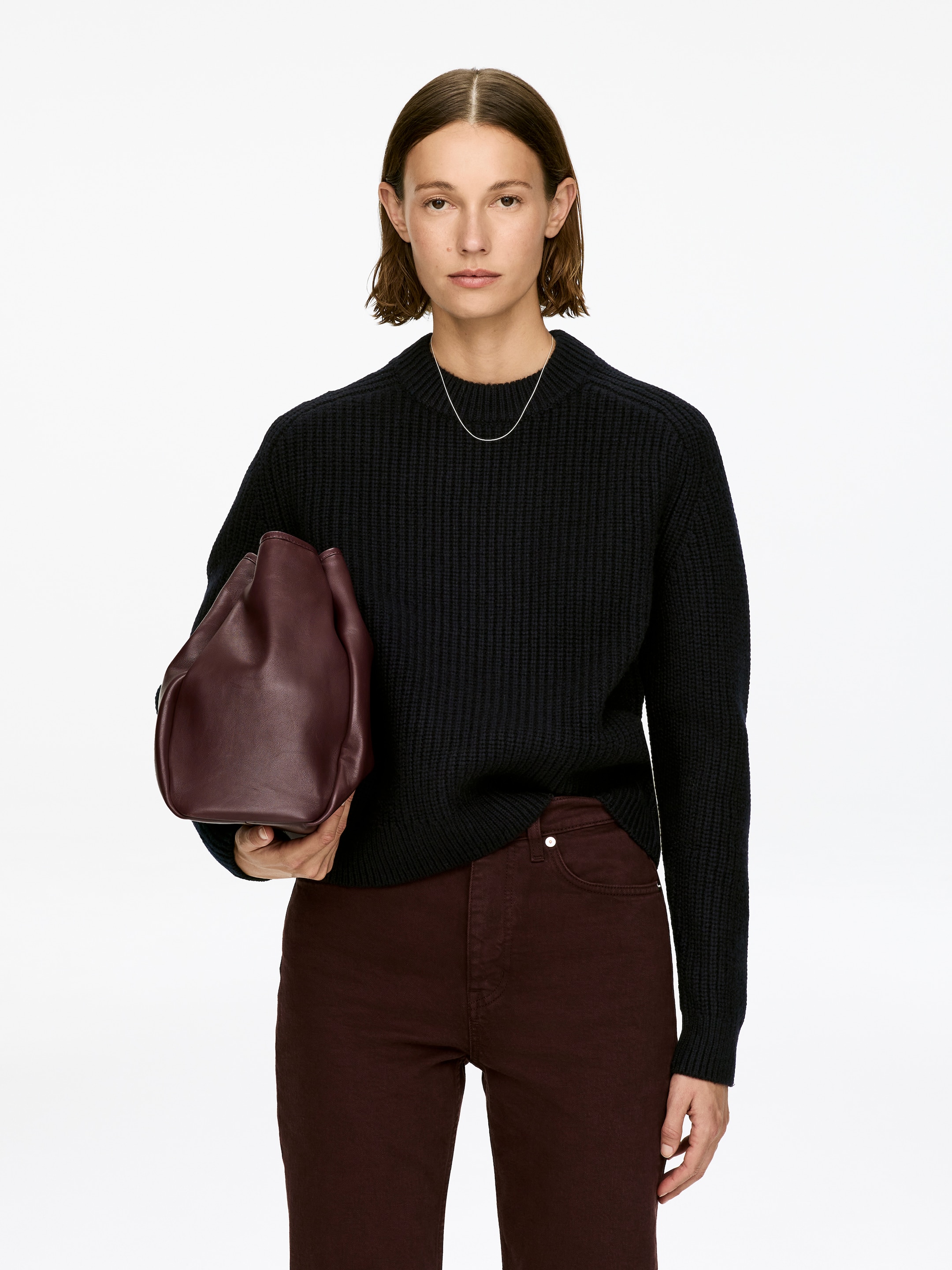 Visa större bild: En svart crew-neck tröja med långa ärmar, med en halv kardiangstickning och ribbade kanter, bärs av modellen. Modellen stylar tröjan med mörkröda byxor och håller i en burgundy väska. Ett tunt silverhalsband är synligt över tröjans halsringning.