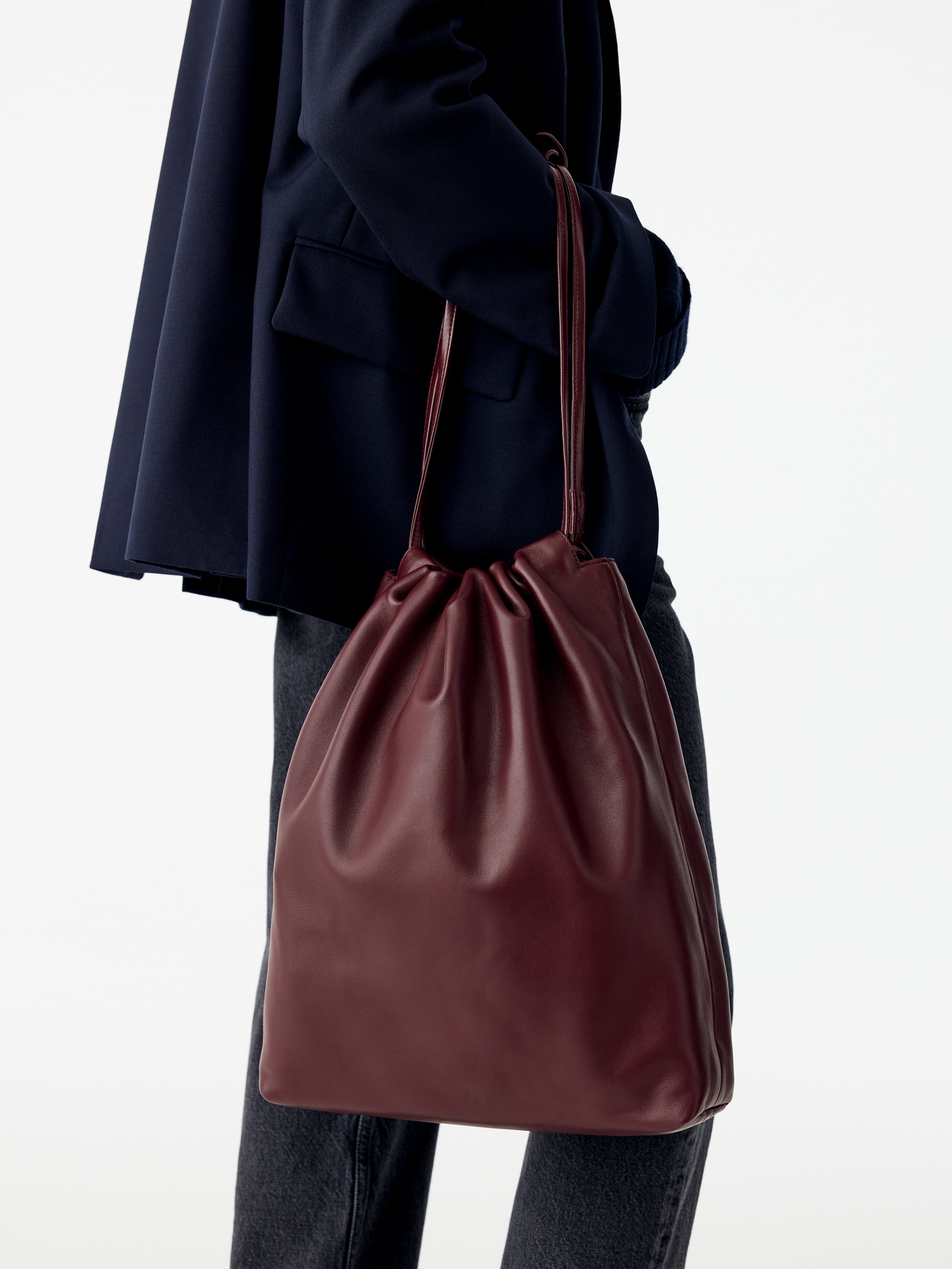 Een diepe bordeauxrode leren tas staat rechtop en toont van voren een nonchalant, op een dustbag geÃ¯nspireerd silhouet. Dunne schouderbanden met trekkoord lopen omhoog vanaf de geplooide bovenopening en zijn samen vastgezet met een bijpassende leren omslag.