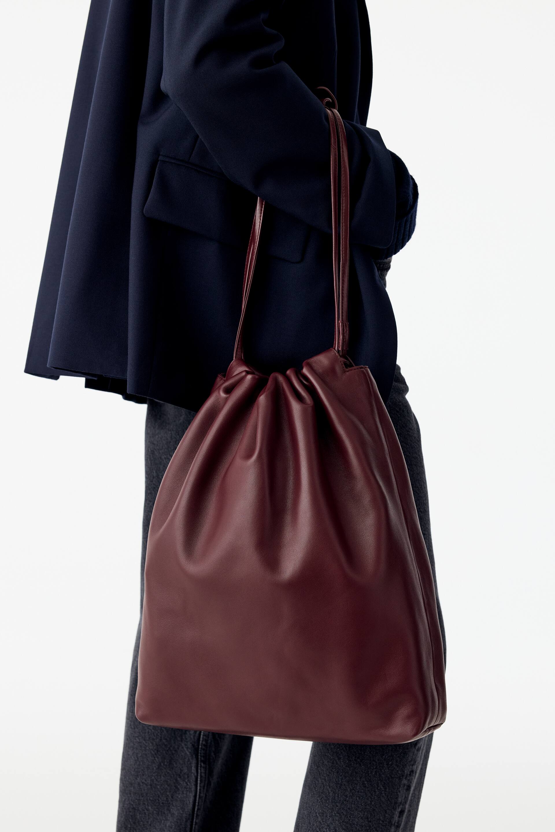 Agrandir l'image: Un sac à cordon en cuir bordeaux, doté d’un haut froncé et de fines bandoulières, est porté à l’épaule par le mannequin. Le sac est associé à un blazer bleu marine foncé et à un jean gris.