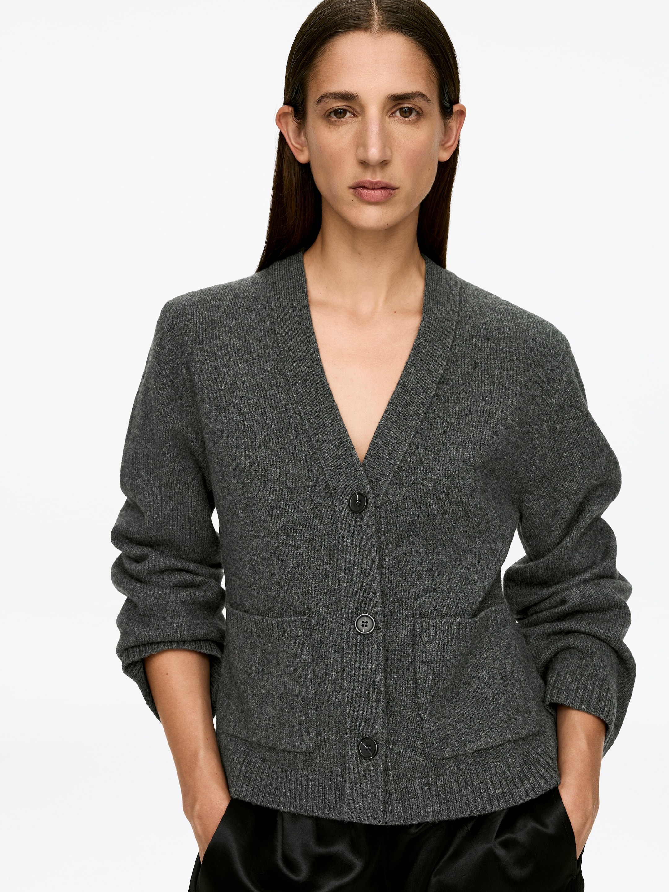 Größeres Bild ansehen: Eine dunkelgraue Strickjacke mit langen Ärmeln, einem V-Ausschnitt, drei dunklen Knöpfen und zwei aufgesetzten Fronttaschen wird von dem Model getragen. Der mittlere Knopf ist geschlossen, die Ärmel sind hochgeschoben und die Hände des Models ruhen in den Fronttaschen.