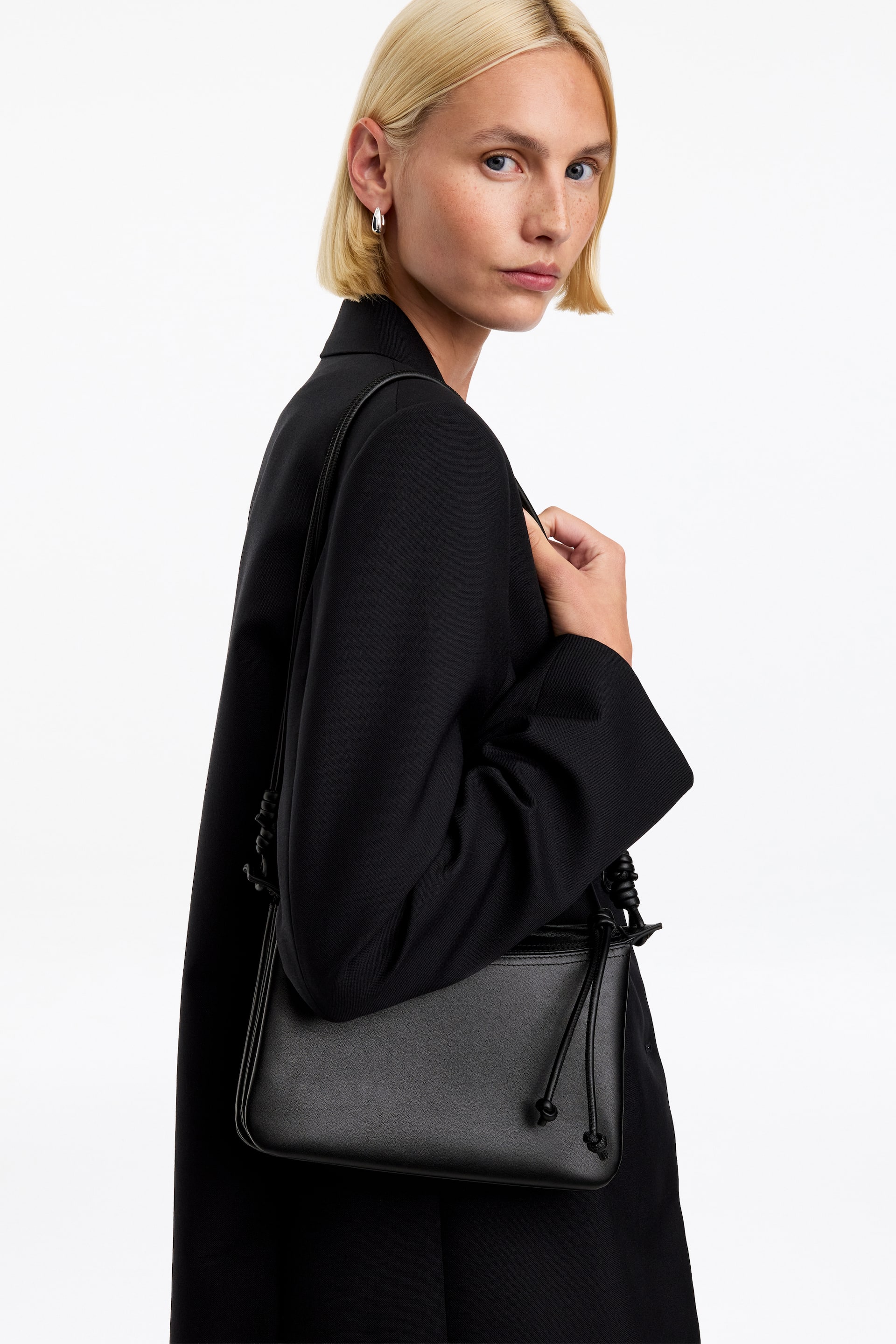 Größeres Bild anzeigen: Eine schwarze Leder-Crossbody-Bag mit strukturierter rechteckiger Form und geknoteten Kordelzug-Details wird vom Model über der Schulter getragen. Das Model hält den dünnen Riemen der Tasche, die über einem schwarzen Blazer gestylt ist.