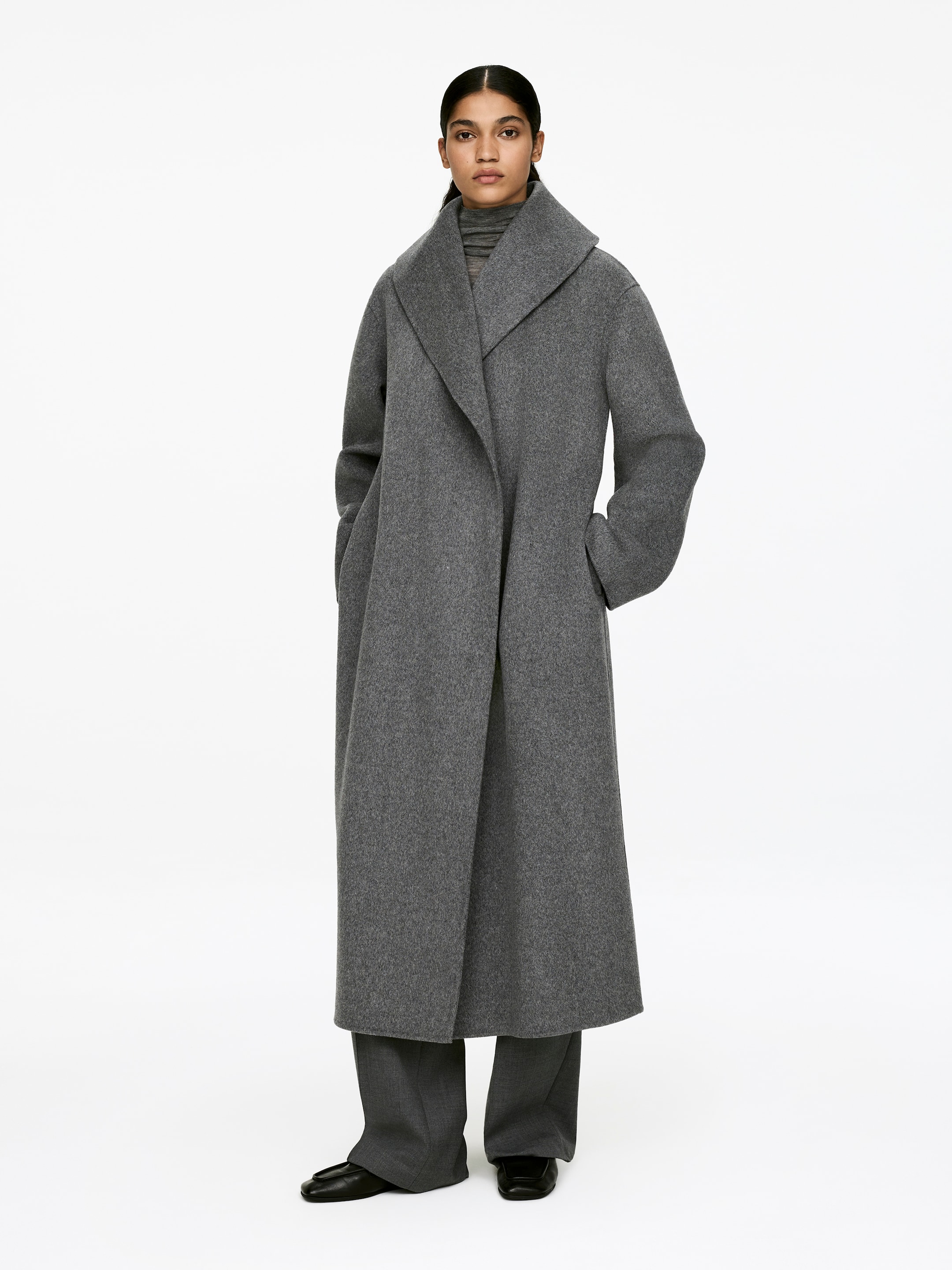 Größeres Bild ansehen: Ein grauer Mantel mit Schalkragen wird von dem Model getragen. Das Model steht mit den Händen in den seitlichen Manteltaschen, die in die Nähte integriert sind. Der lange Mantel, aus einem schweren grauen Stoff gefertigt, wird über einem grauen Rollkragenpullover und grauen Hosen getragen.