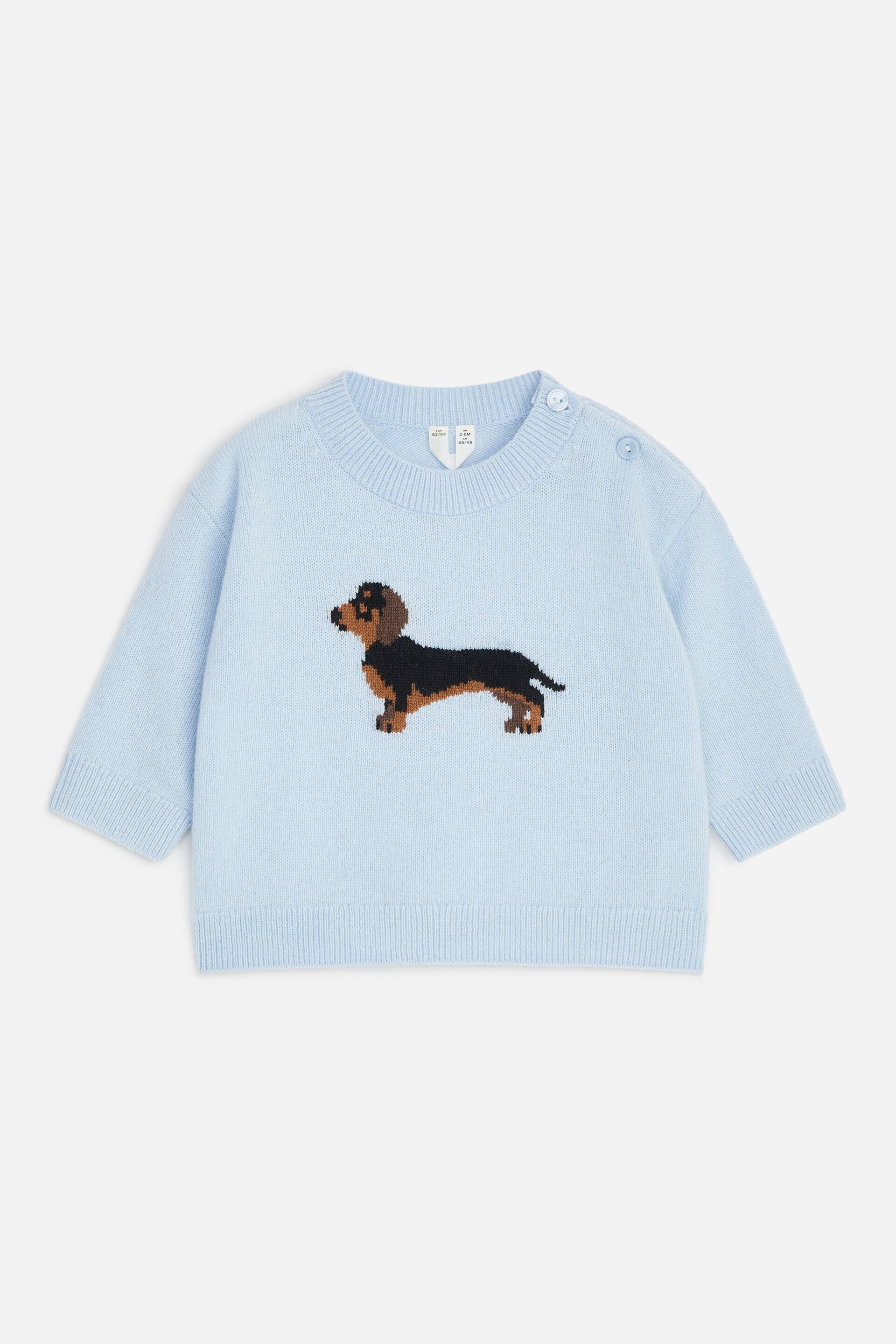 View larger image: A light blue knitted baby jumper is shown from the front, featuring a knitted black and tan dachshund design in the centre. The jumper includes a ribbed crew neck with two button fastenings on the left shoulder, along with ribbed cuffs and a ribbed hem.
