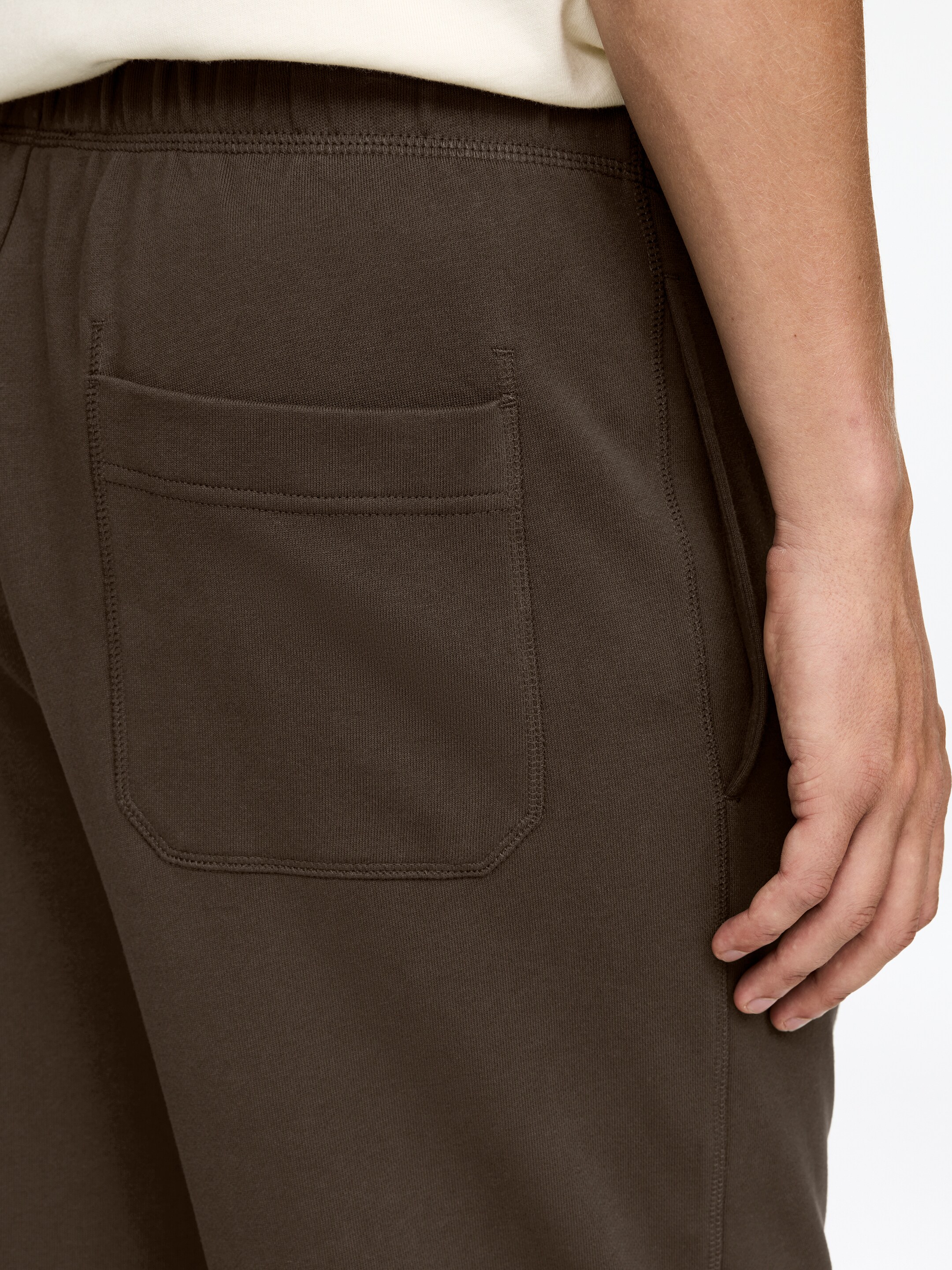 Ingrandisci l'immagine: I pantaloni della tuta marroni sono mostrati da un'angolazione posteriore, caratterizzati da una cintura elasticizzata e una tasca arrotondata sul posteriore sinistro. Un cordino è visibile lungo la cucitura laterale.