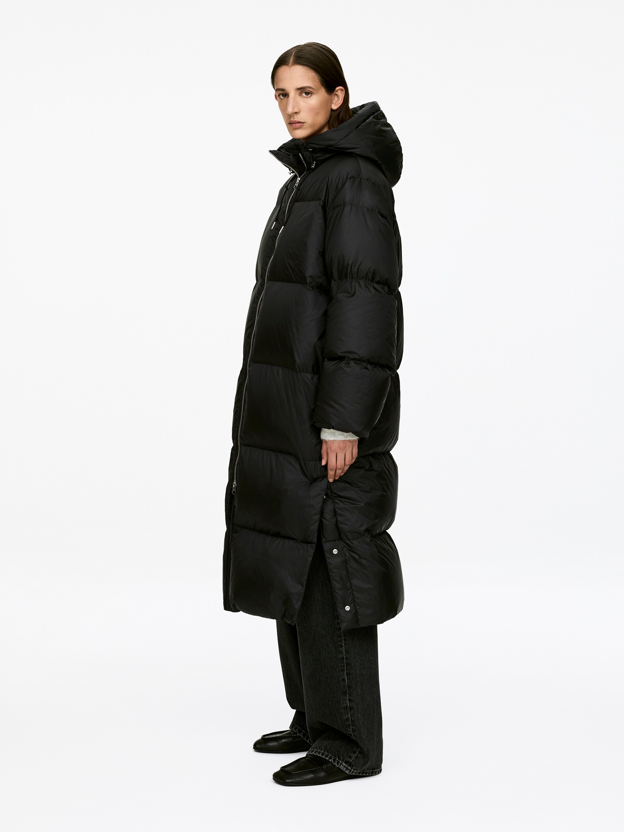 Visa större bild: Upcycled Down Long Coat bärs av modellen. Den svarta, huvtäckta pufferjackan har horisontell baffling, hög krage och en tvåvägs dragkedja framtill, och den är ankellång med sidoventiler vid fållen som är öppnade.