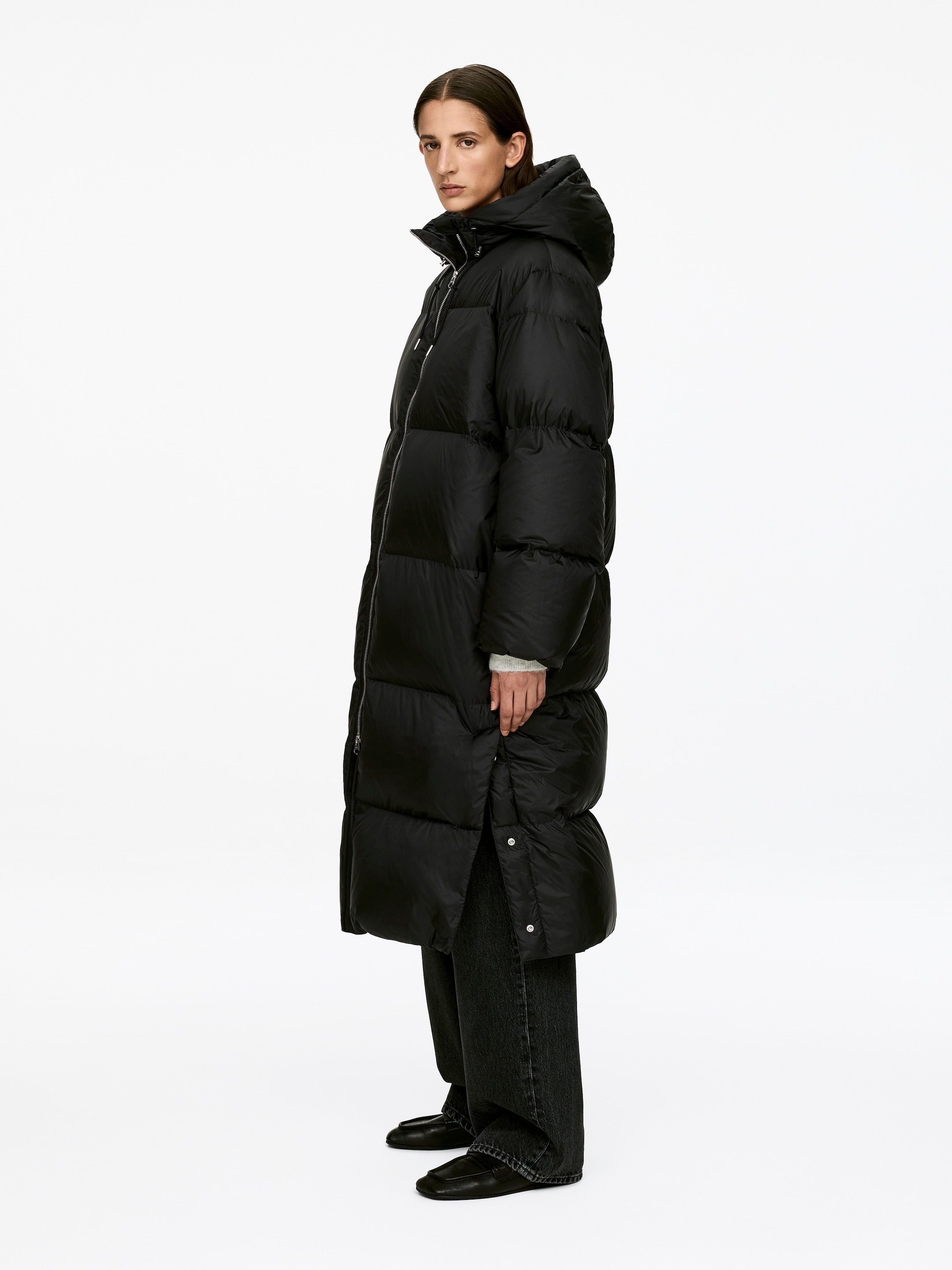 View larger image: Upcycled Down Long Coat is being worn by the model. The black, hooded puffer coat features horizontal baffling, a high collar, and a two-way front zipper, and it is ankle-length with side vents at the hem being unfastened.