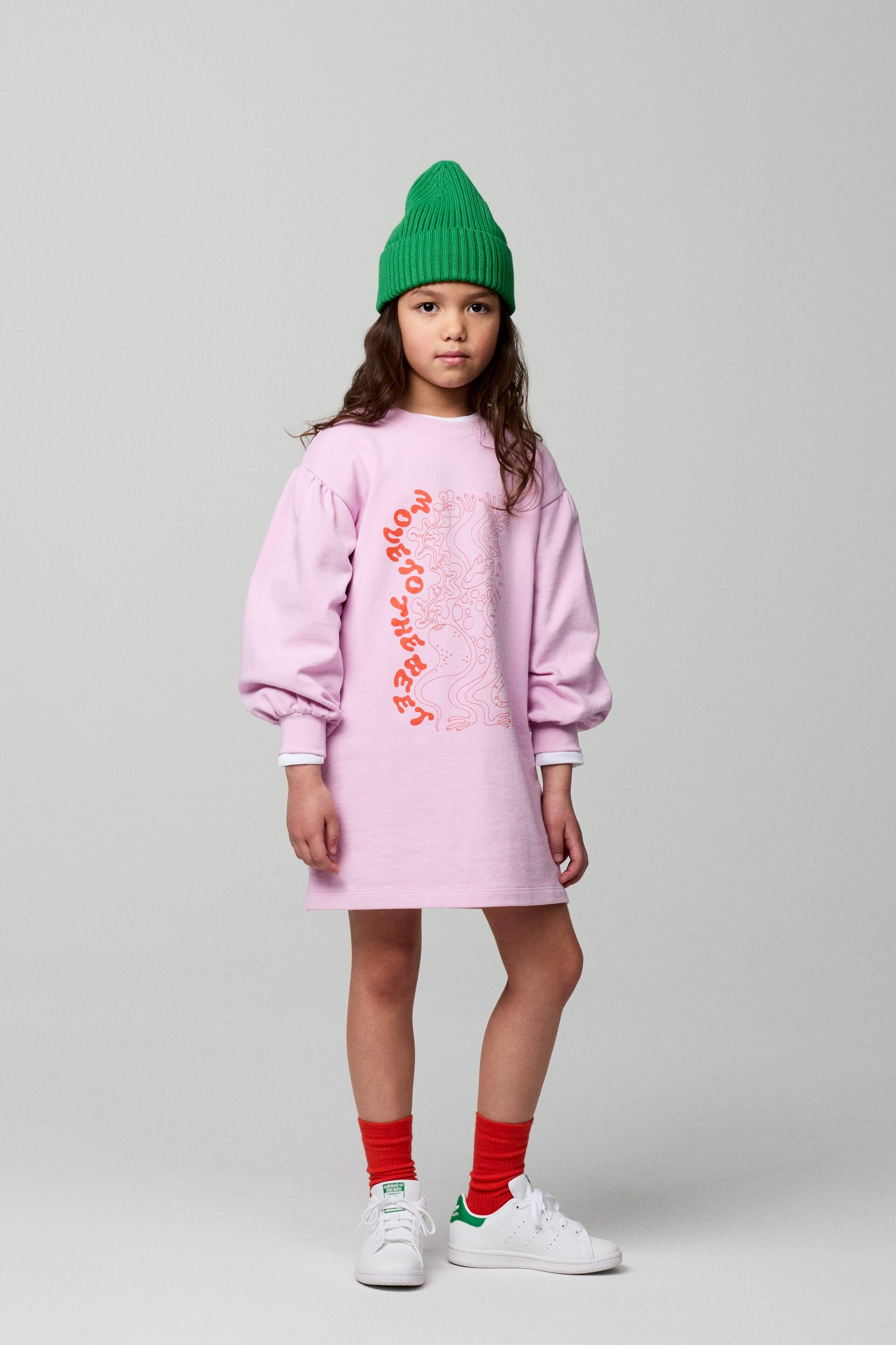 Agrandir l'image: Une robe sweat-shirt rose clair, présentant une coupe ample et tombant au-dessus du genou, est dotée de manches longues et bouffantes avec des poignets côtelés révélant une couche blanche en dessous. Un grand imprimé graphique textuel et abstrait orange et rouge recouvre le devant de la robe, et un bonnet en maille côtelée vert est également présenté.