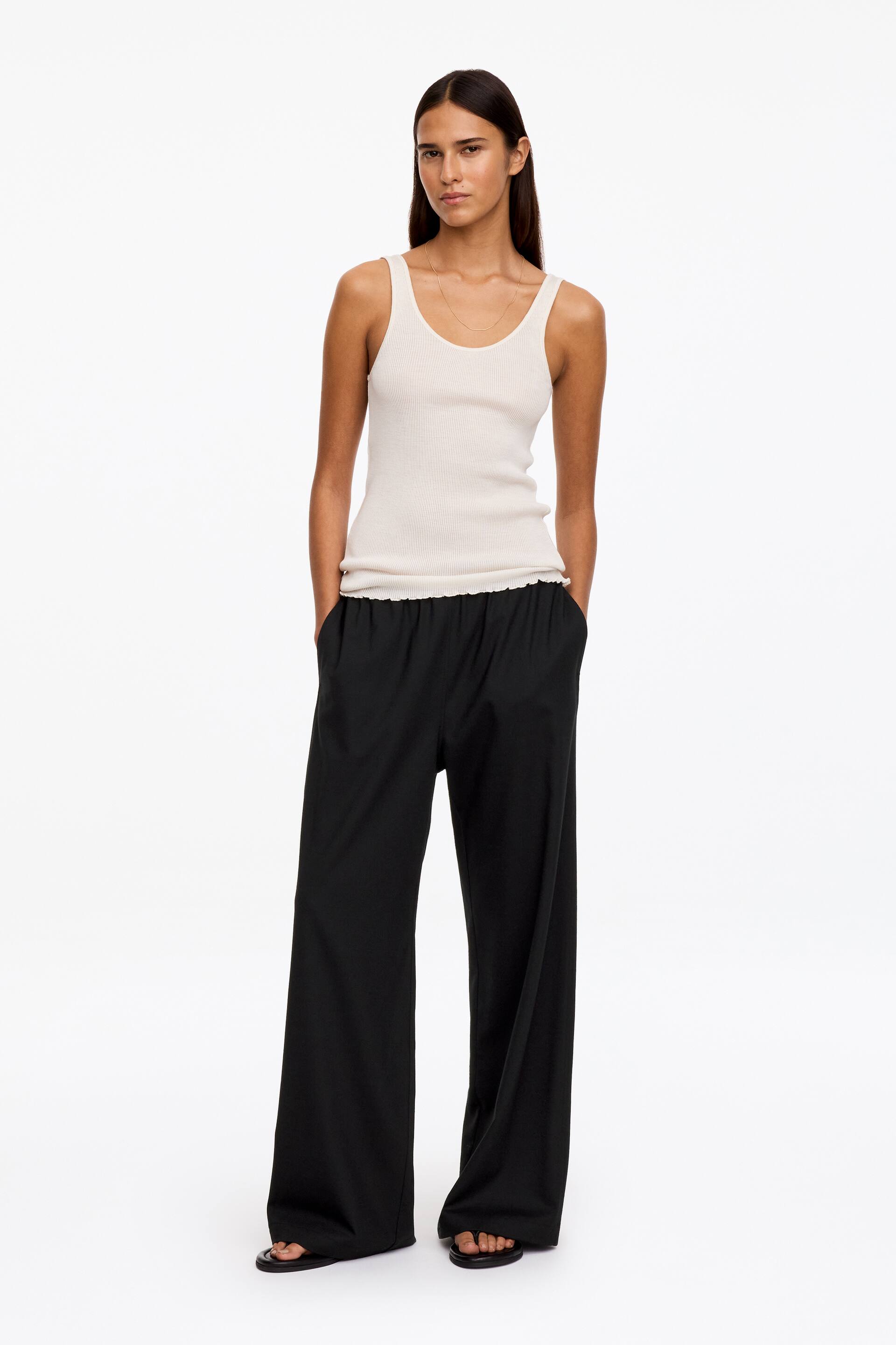 Ver imagen más grande: La modelo lleva pantalones negros de pernera ancha con cintura elástica y está de pie con las manos apoyadas en los bolsillos laterales. Los pantalones de corte relajado se combinan con un top de tirantes de canalé en color crema y sandalias planas negras.
