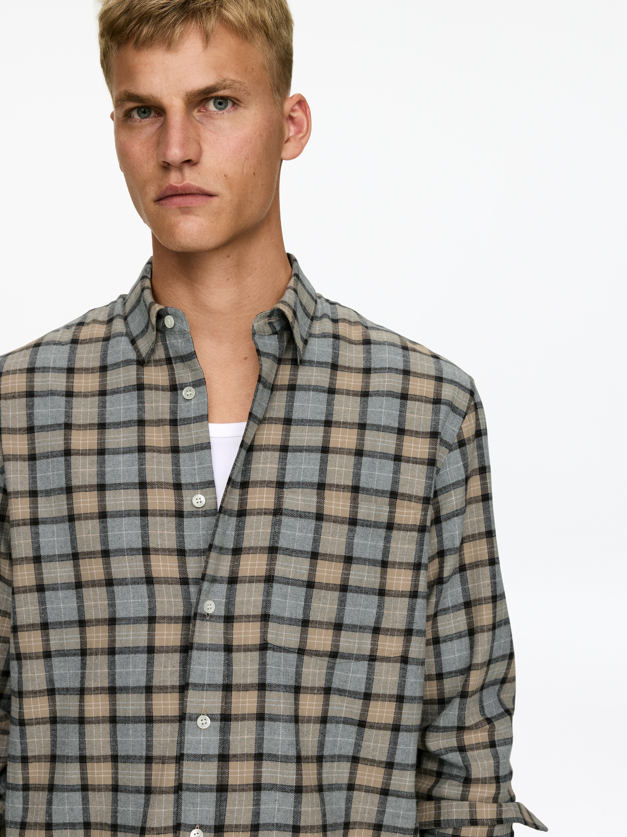 Ver imagen más grande: Una camisa de cuadros se muestra desde el frente, con un patrón de cuadros grises, marrones y negros con líneas blancas finas. Su cuello puntiagudo está abierto, y la tapeta delantera está parcialmente desabrochada, revelando una camiseta blanca debajo. Un único bolsillo en el pecho izquierdo es visible, y una de las mangas largas muestra un puño enrollado.