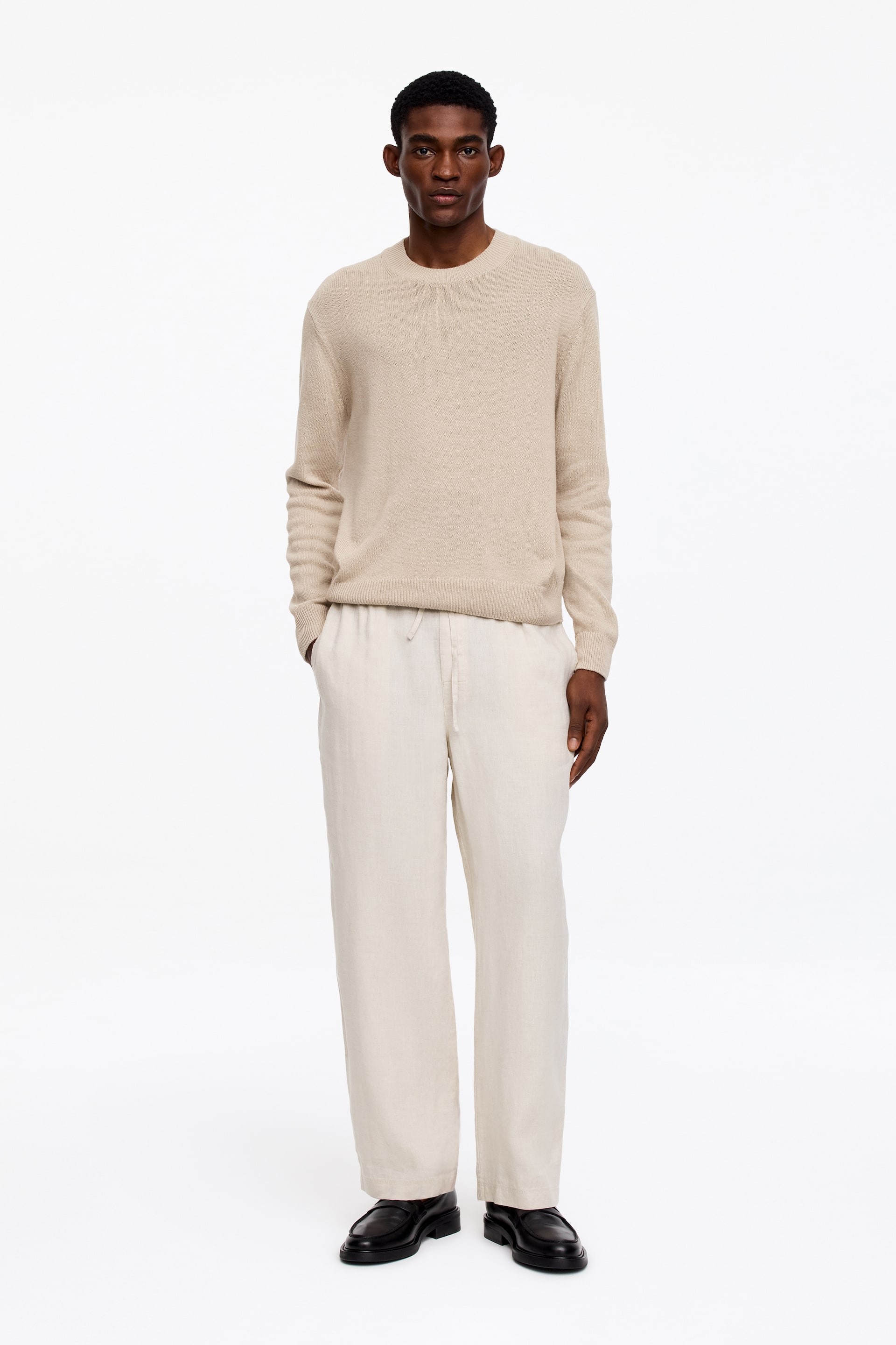 Visa större bild: En beige crewneck-tröja med ribbade muddar och fåll bärs, vilket visar en stickad textur. Den kombineras med krämfärgade, vida byxor med synligt dragsnöre i midjan och ett texturerat tyg, med svarta loafers med kraftiga sulor som fullbordar looken.