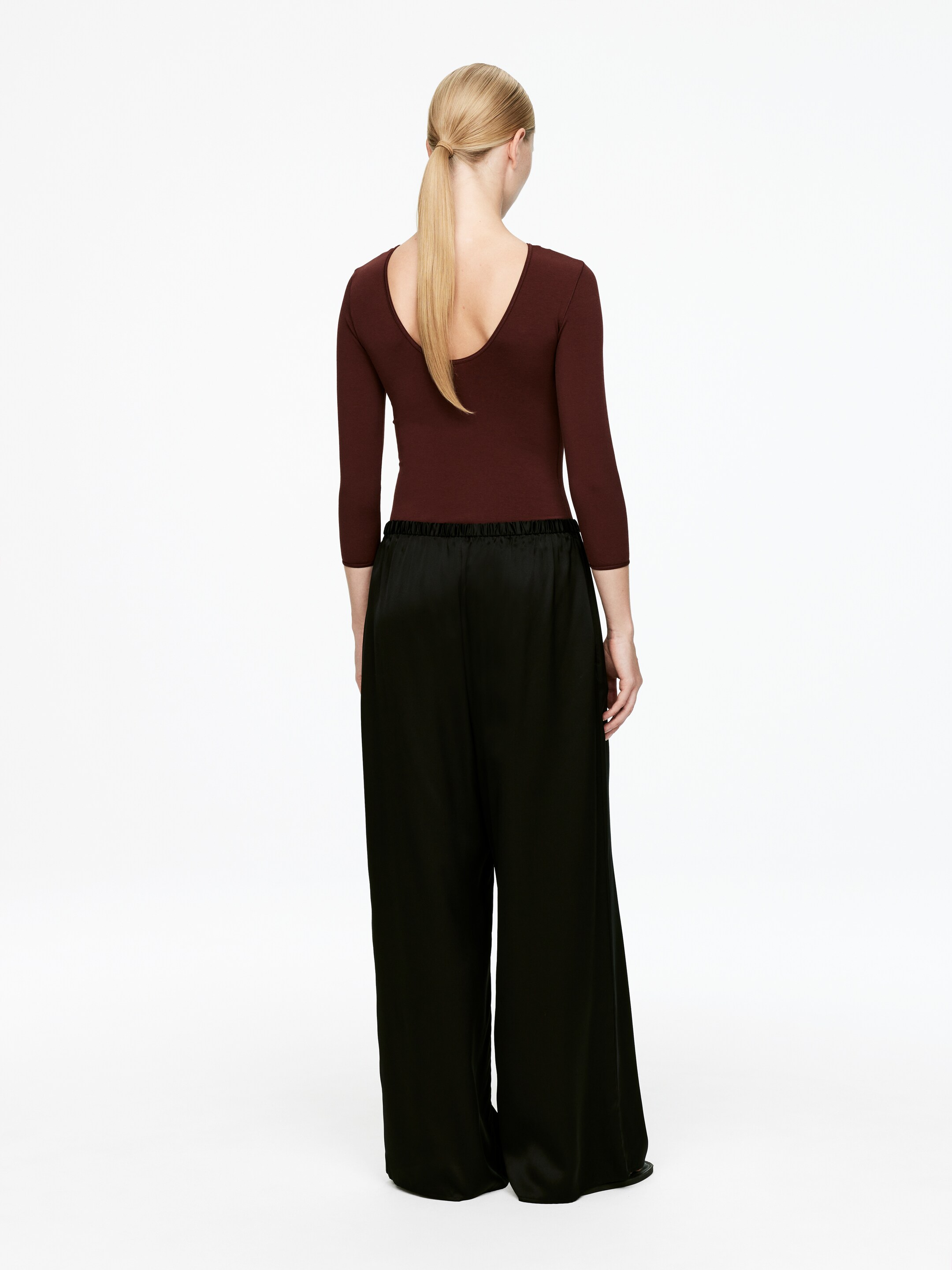 Ver imagen más grande: Una blusa de color marrón rojizo oscuro con un escote profundo en la espalda y mangas tres cuartos se lleva puesta, vista desde atrás, con pantalones negros de pierna ancha confeccionados con una tela lustrosa, que se acumulan en el dobladillo.