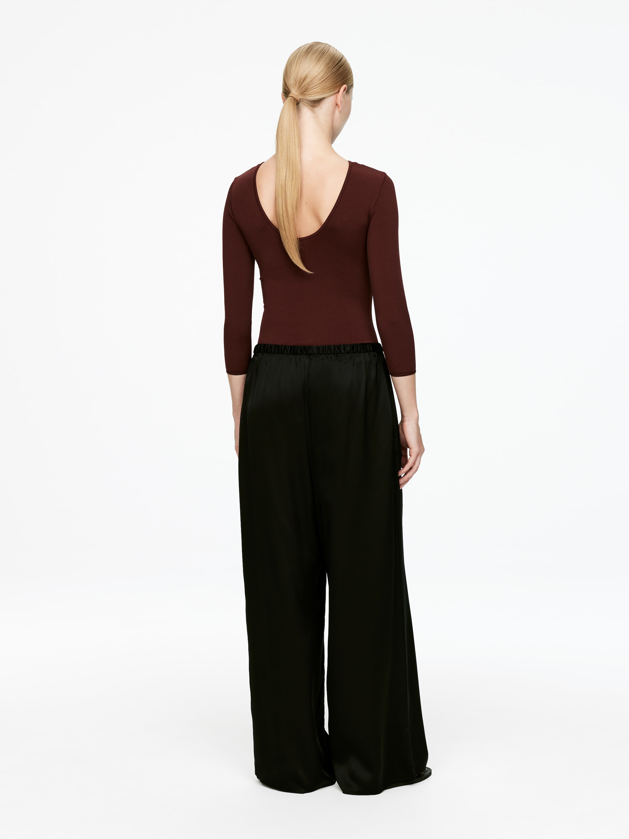 Ver imagen más grande: Una blusa de color marrón rojizo oscuro con un escote profundo en la espalda y mangas tres cuartos se lleva puesta, vista desde atrás, con pantalones negros de pierna ancha confeccionados con una tela lustrosa, que se acumulan en el dobladillo.