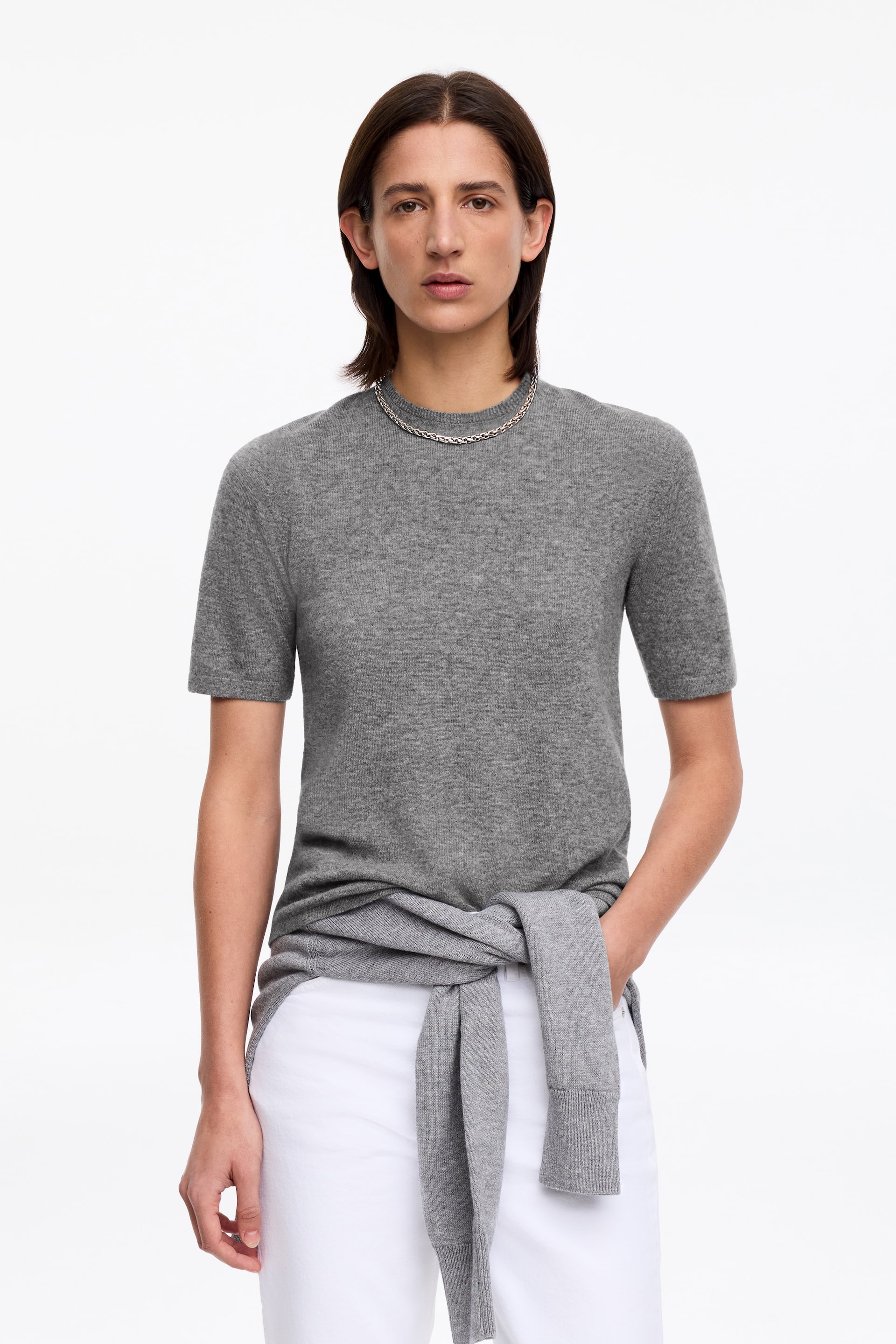 Ingrandisci l'immagine: Un maglione grigio a maniche corte viene presentato frontalmente, caratterizzato da uno scollo rotondo e una texture a maglia fine, con un altro maglione grigio abbinato legato in vita.