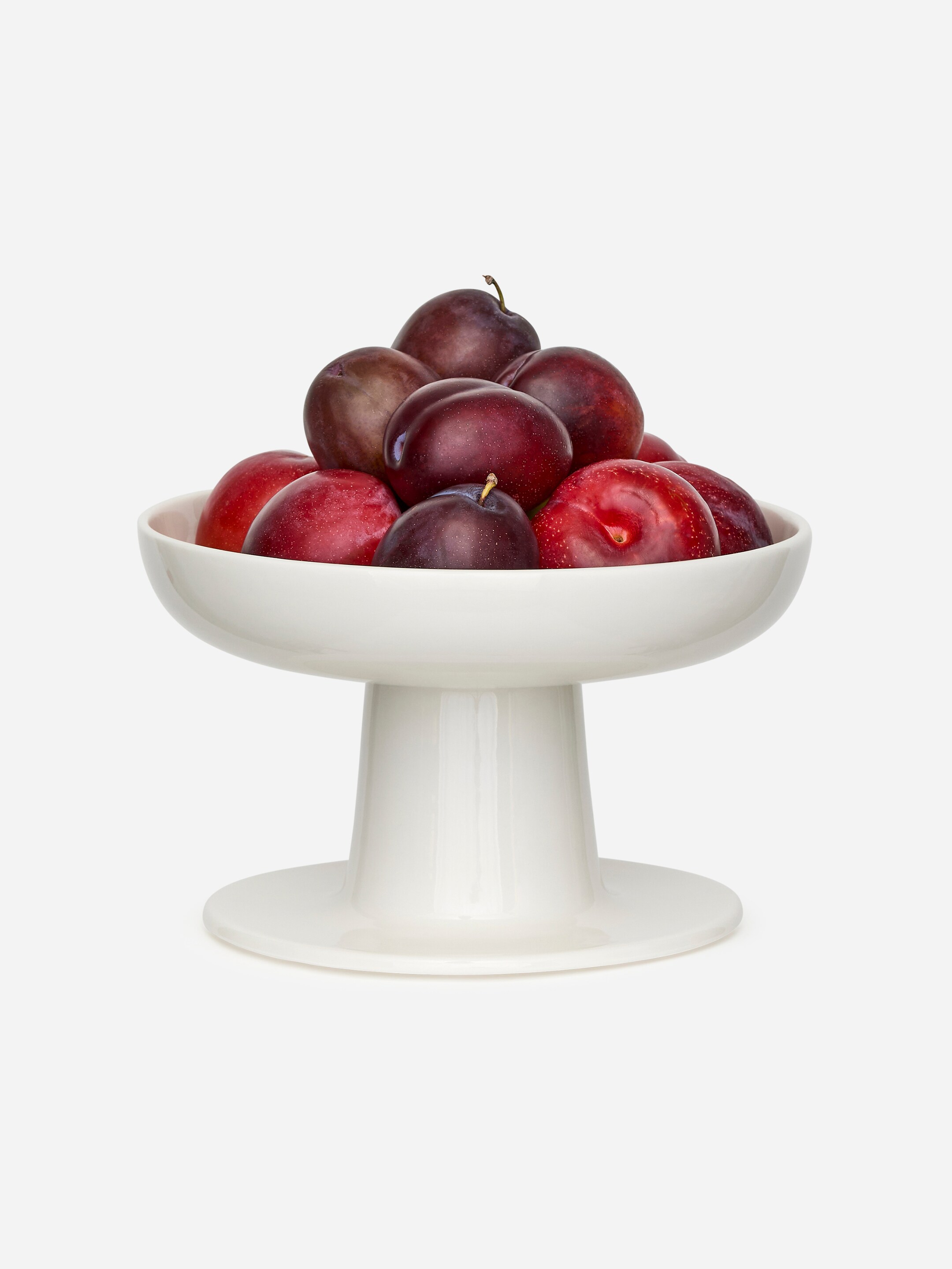 Visa större bild: En vit, glaserad stengods serveringsskål presenteras FRAMIFRÅN, med en höjd på 14 cm och en diameter på 22 cm. Den breda, grunda skålen vilar på en tjock, utsvängd piedestalbas och håller för närvarande en hög med plommon.