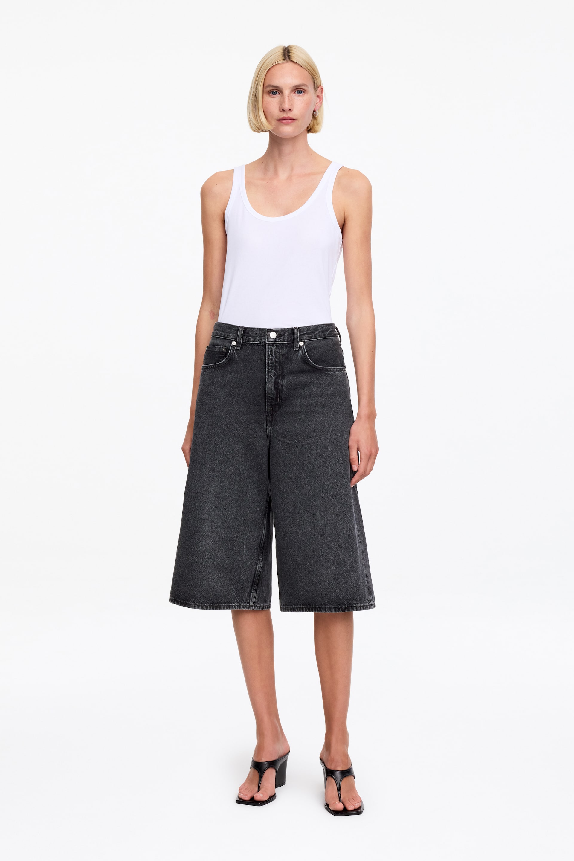 Se større billede: En hvid tanktop præsenteres, med en rund halsudskæring og brede skulderstropper, og bæres nede i mørkvaskede shorts.