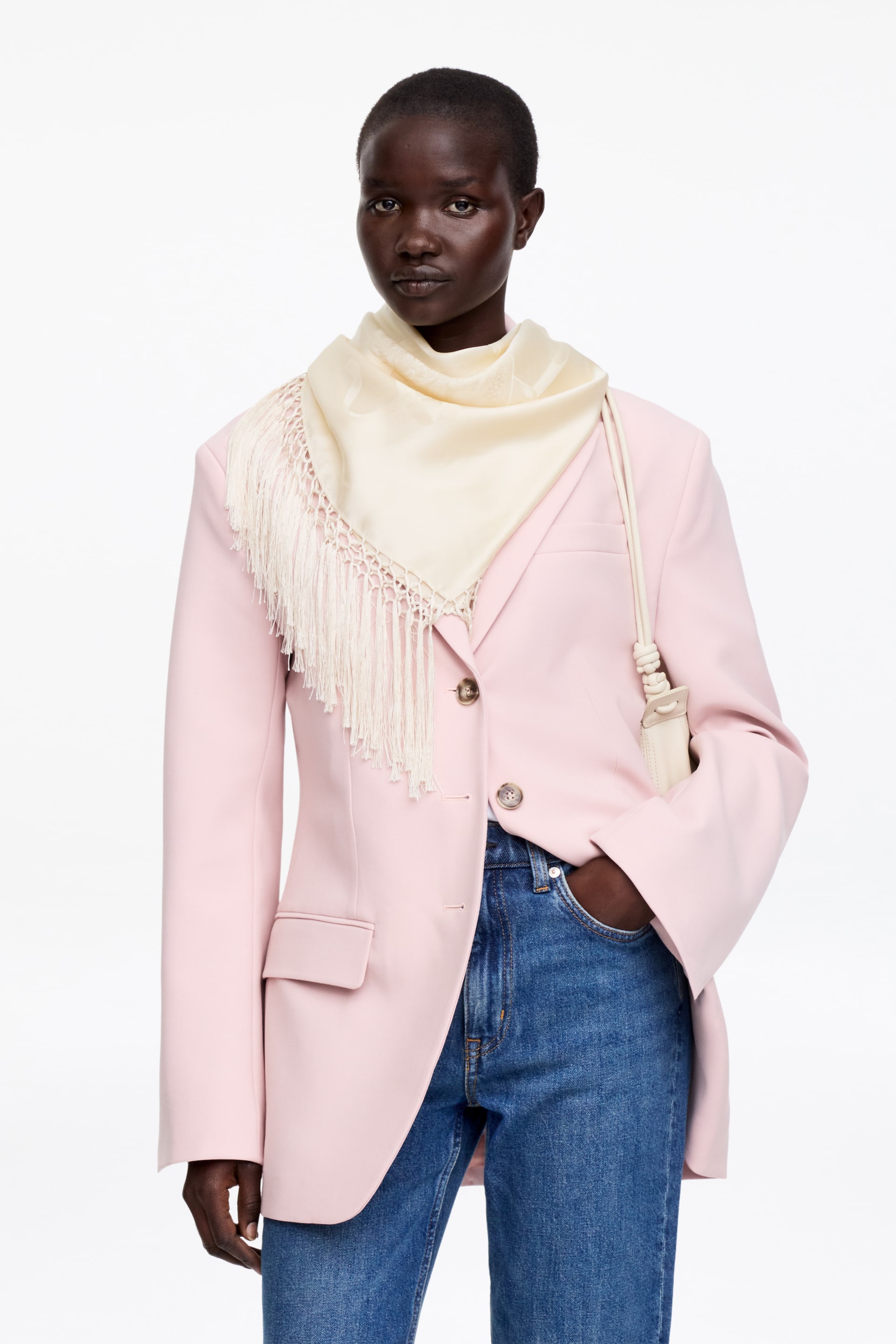 Visualizza immagine più grande: Un blazer monopetto rosa chiaro, mostrato da una vista frontale leggermente angolata e caratterizzato da due bottoni marroni e tasche a pattina, è indossato sopra jeans blu a gamba dritta con lavaggio medio. Una sciarpa triangolare color crema con bordi sfrangiati è drappeggiata sulla spalla sinistra, mentre una piccola borsa a tracolla color crema con tracolla annodata è appesa alla spalla destra.