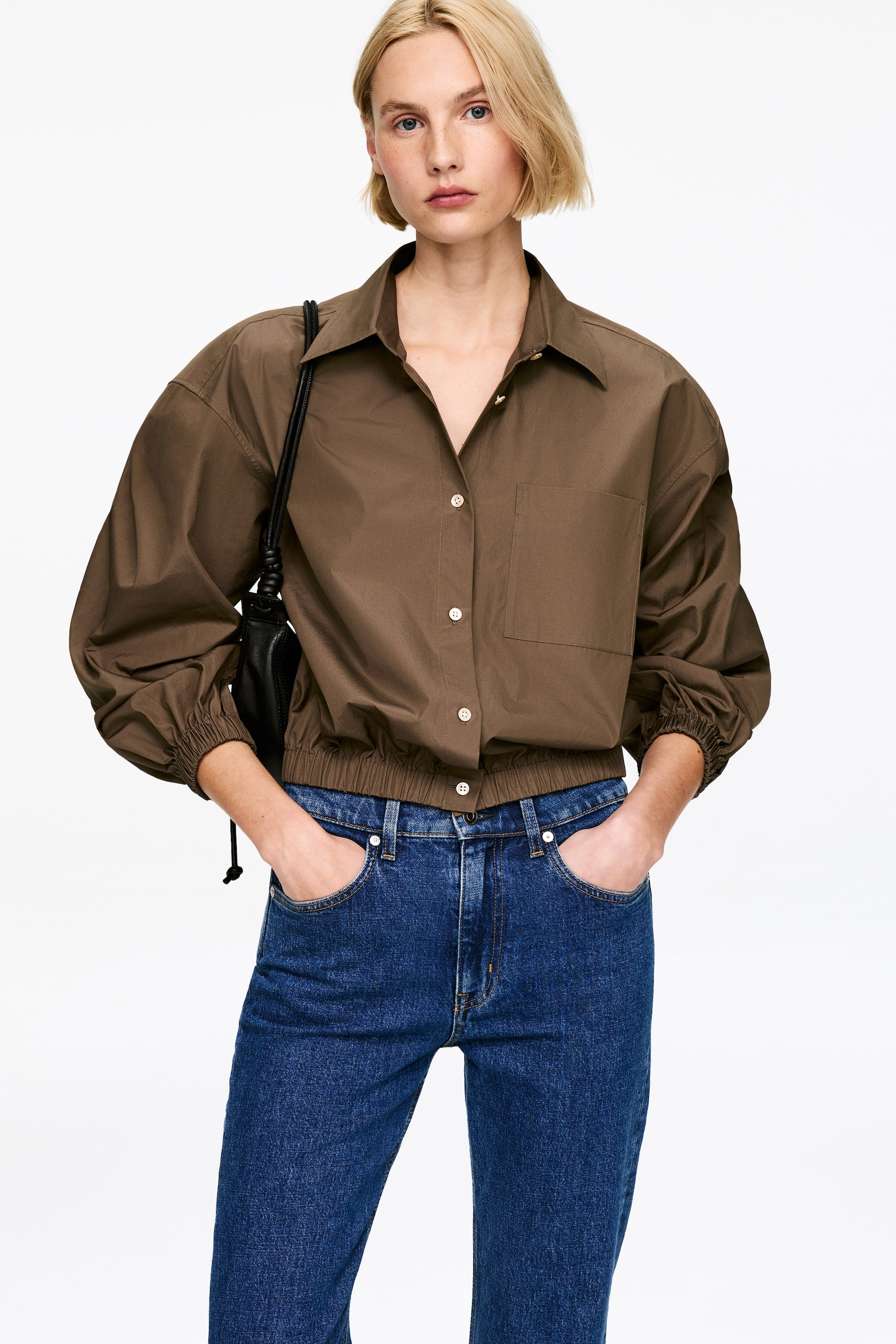 Grösseres Bild ansehen: Ein khakifarbenes Hemd wird von vorne präsentiert, mit einem spitzen Kragen, einem Knopfverschluss mit hellen Knöpfen und einer aufgesetzten Brusttasche auf der linken Seite. Es ist mit voluminösen langen Ärmeln gestaltet, die in elastischen Bündchen enden, sowie einem elastischen Saum, der die Taille betont.