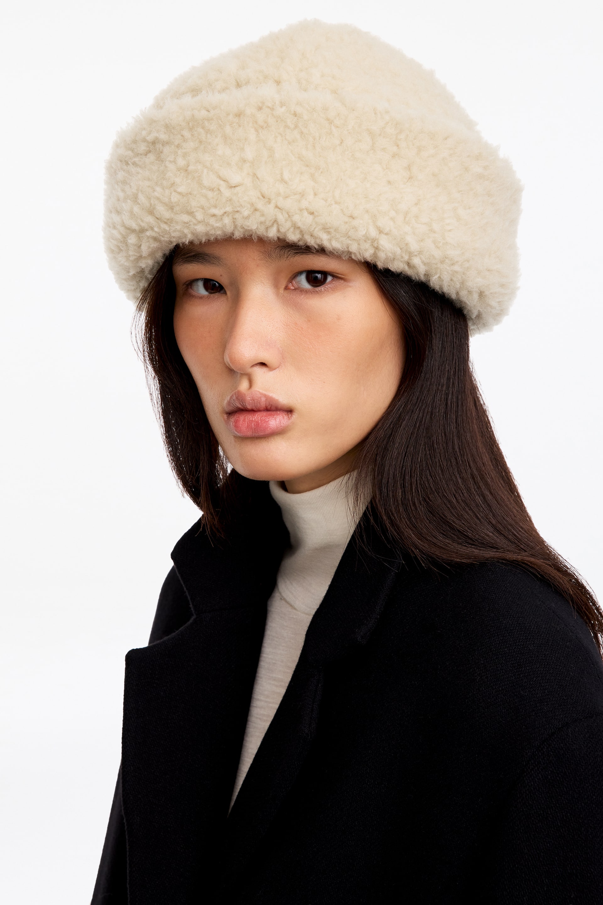 Visualizza l'immagine ingrandita: Il modello indossa un cappello a pelo ripiegabile color crema, realizzato in tessuto di lana testurizzato e riccio. Il cappello presenta una tesa ripiegata spessa e arrotondata ed è abbinato a un dolcevita color crema e a un cappotto nero.