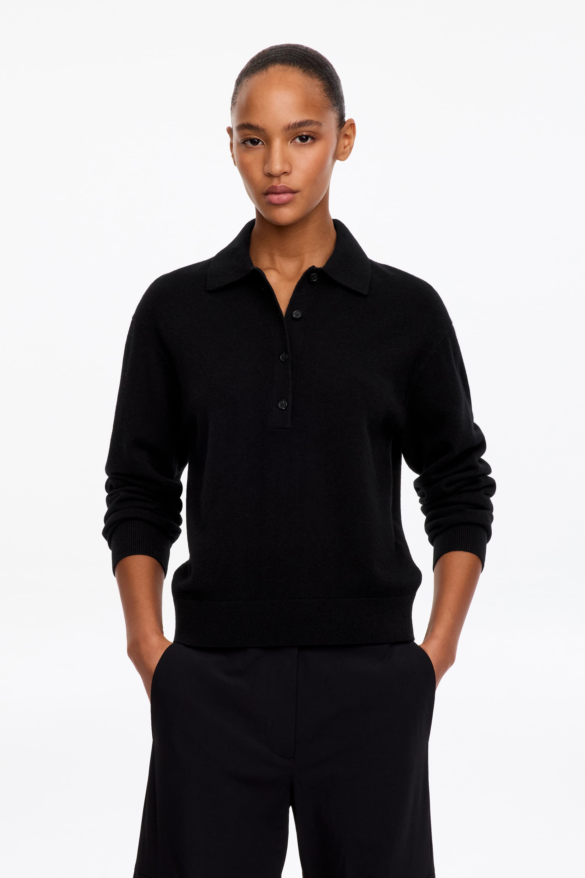 Visualizza l'immagine ingrandita: Una polo nera in maglia con colletto classico, abbottonatura parziale e bordi a coste è indossata dalla modella. Il top a maniche lunghe è abbinato a pantaloni neri coordinati.