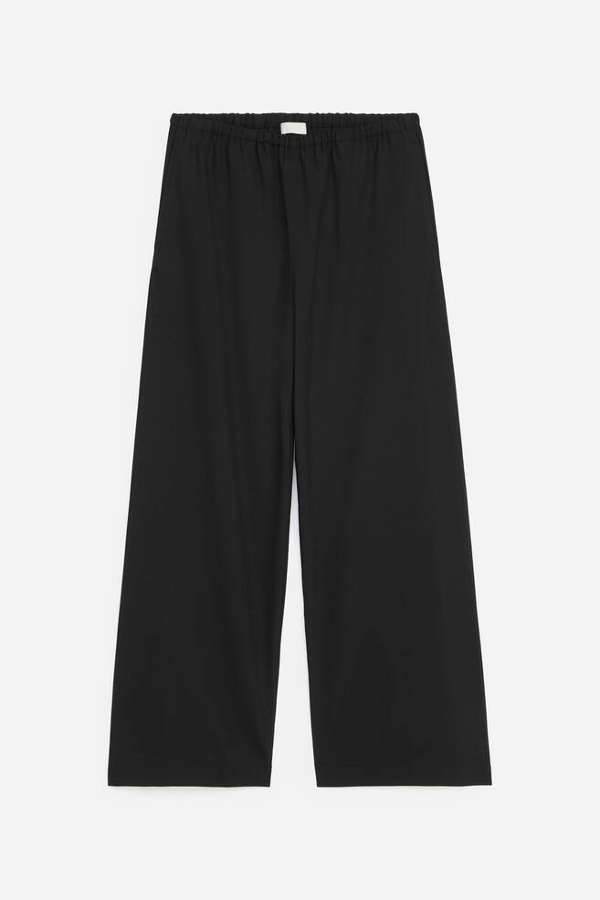Eine schwarze Wide-Leg-Hose ist von vorne zu sehen und verfÃ¼gt Ã¼ber einen gerafften, elastischen Bund sowie eine lockere, gerade Silhouette. Die Hose ist flach liegend prÃ¤sentiert und zeigt ihr weiches, matt strukturiertes Material sowie sauber gearbeitete, schlichte SÃ¤ume. - 1