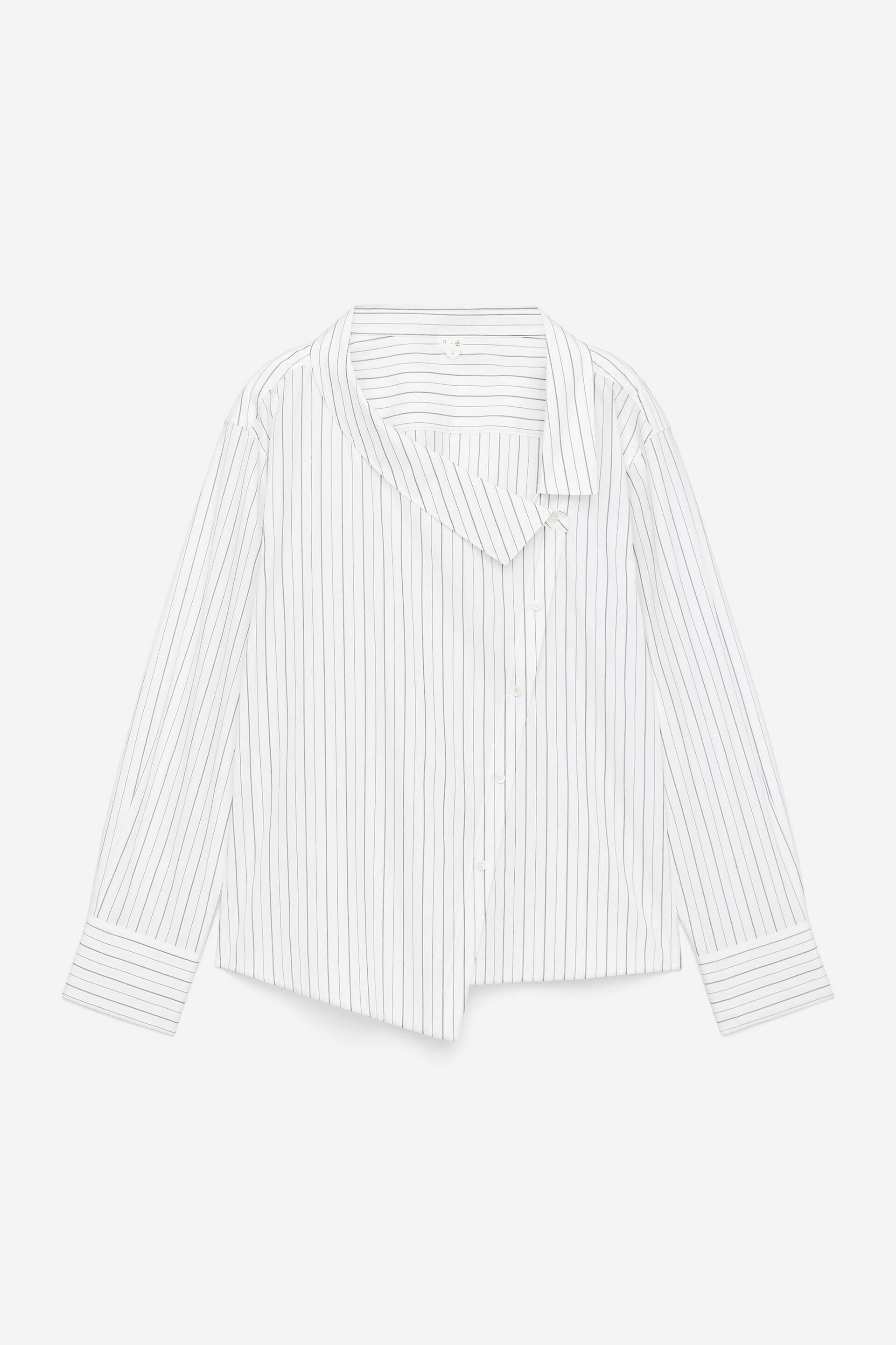 Vis større bilde: En hvit skjorte med tynne mørke vertikale striper blir presentert fra forsiden, og viser en åpen, asymmetrisk krage med et overlappende frontpanel. Den har også en delvis knappestolpe, lange ermer med brede mansjetter, og en vinklet, asymmetrisk kant.
