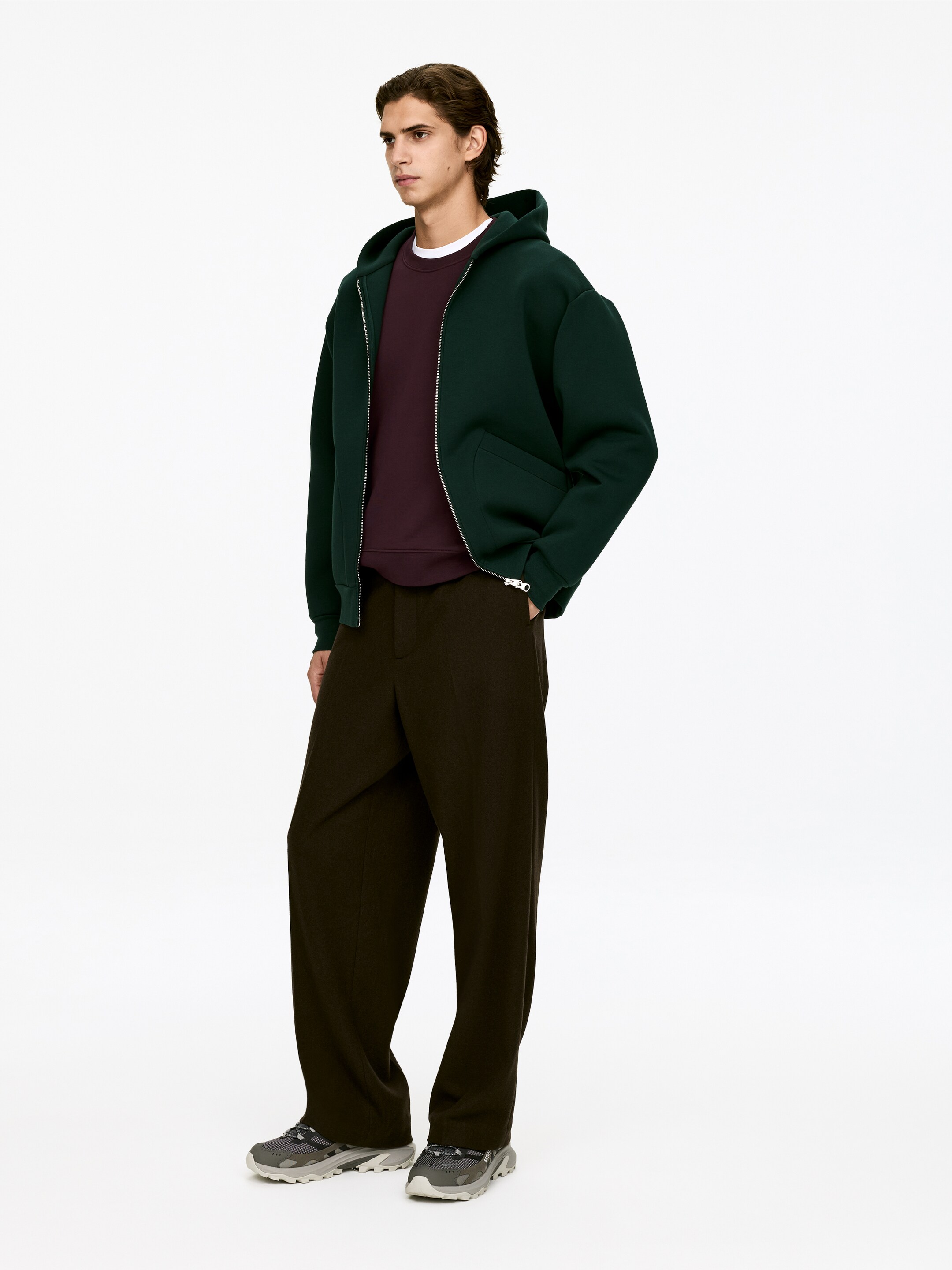 Ver imagen más grande: Una chaqueta con capucha de color verde oscuro, con cremallera completa y de gran tamaño, se presenta en una vista de tres cuartos, mostrando un cierre de cremallera plateado y bolsillos frontales. Está combinada con un jersey de cuello redondo burdeos y una camiseta blanca, con pantalones de pierna ancha de color marrón oscuro y zapatillas deportivas gris y negras que completan el conjunto.