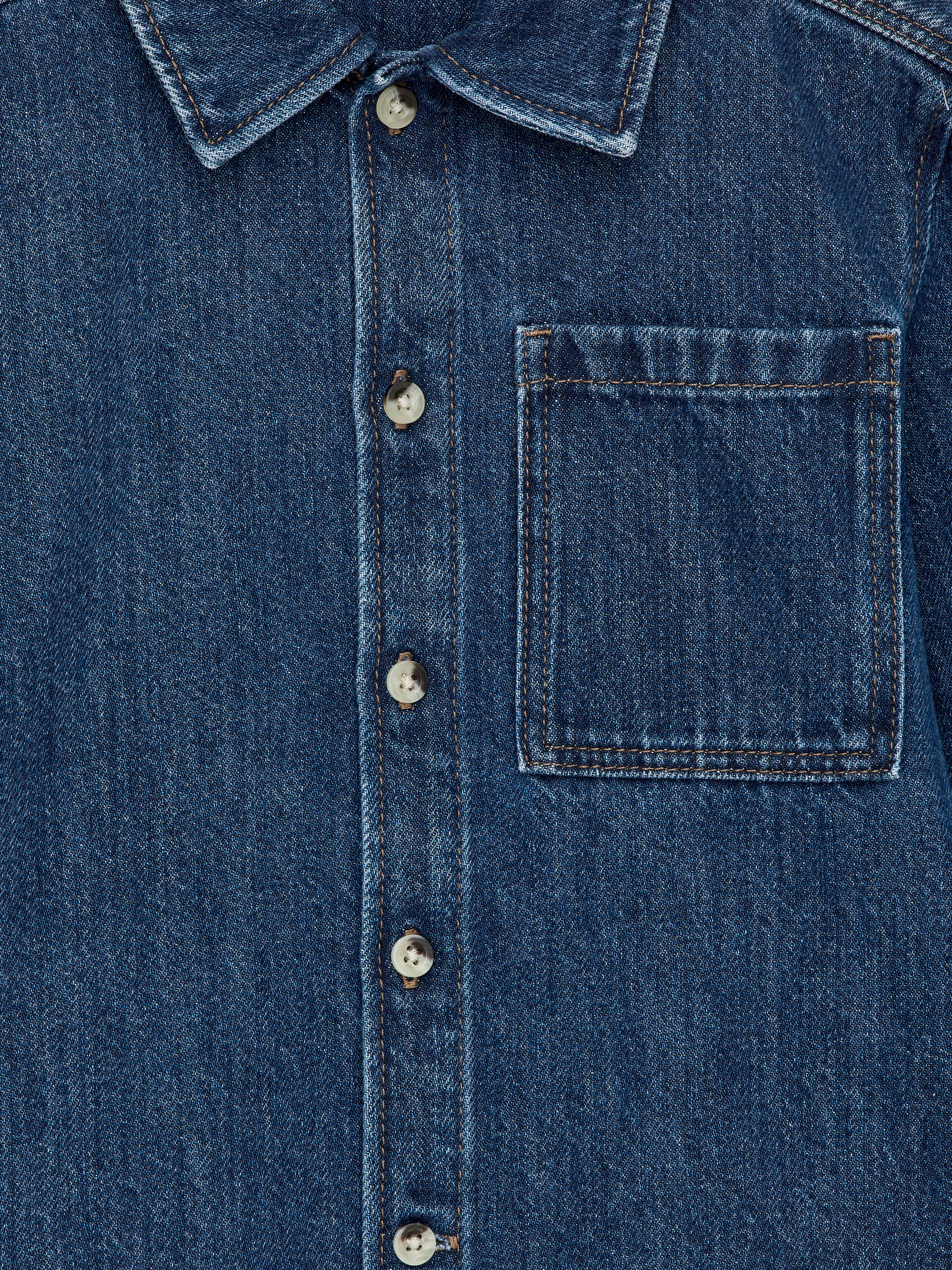 Ingrandisci l'immagine: Una camicia di jeans blu è visibile in una qualità leggermente più pesante con una finitura lavata. Presenta una chiusura frontale con bottoni di colore chiaro, un colletto a punta e una singola tasca applicata sul petto destro, con cuciture a contrasto che dettagliano le cuciture.