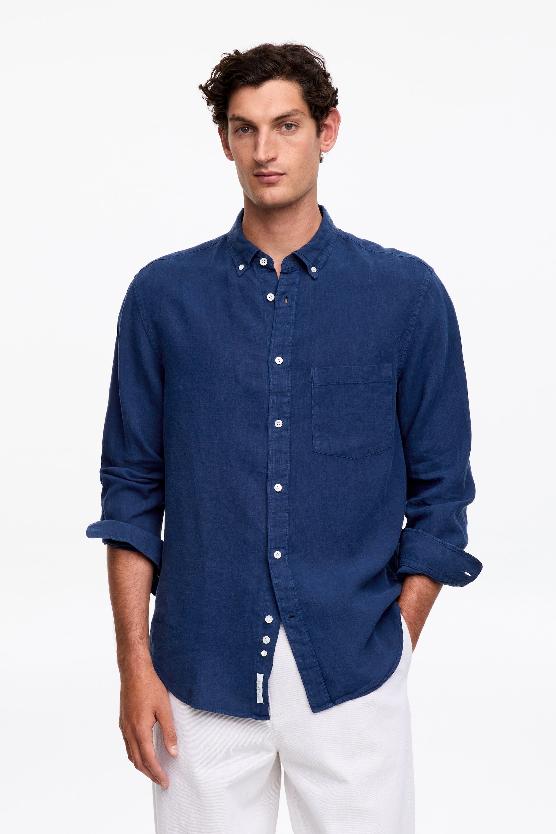 Ingrandisci l'immagine: Una camicia in lino blu scuro con colletto abbottonato è presentata frontalmente, caratterizzata da un colletto button-down, bottoni bianchi sulla pattina anteriore e una tasca applicata sul petto sinistro. Le maniche lunghe sono arrotolate e l'orlo curvo è indossato fuori dai pantaloni.