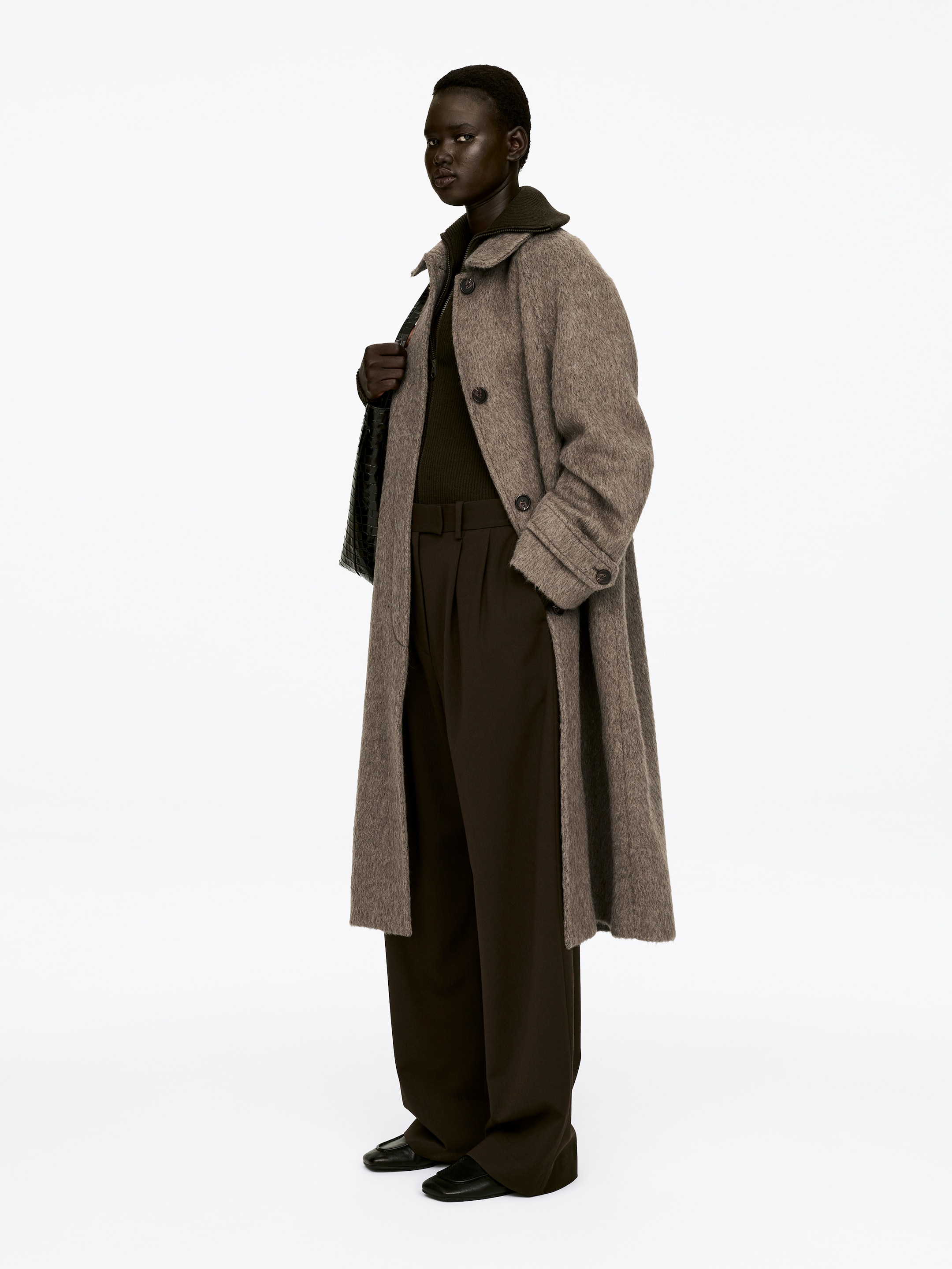 View larger image: A long, single-breasted coat in a brushed heathered brown wool and alpaca blend is being worn by the model. The coat features a spread collar, raglan sleeves, visible front buttons, welt pockets, and buttoned tabs at the cuffs. The model's hand is holding the coat's lapel, partially revealing a dark ribbed top layered underneath, while a dark bag is being carried on the shoulder.