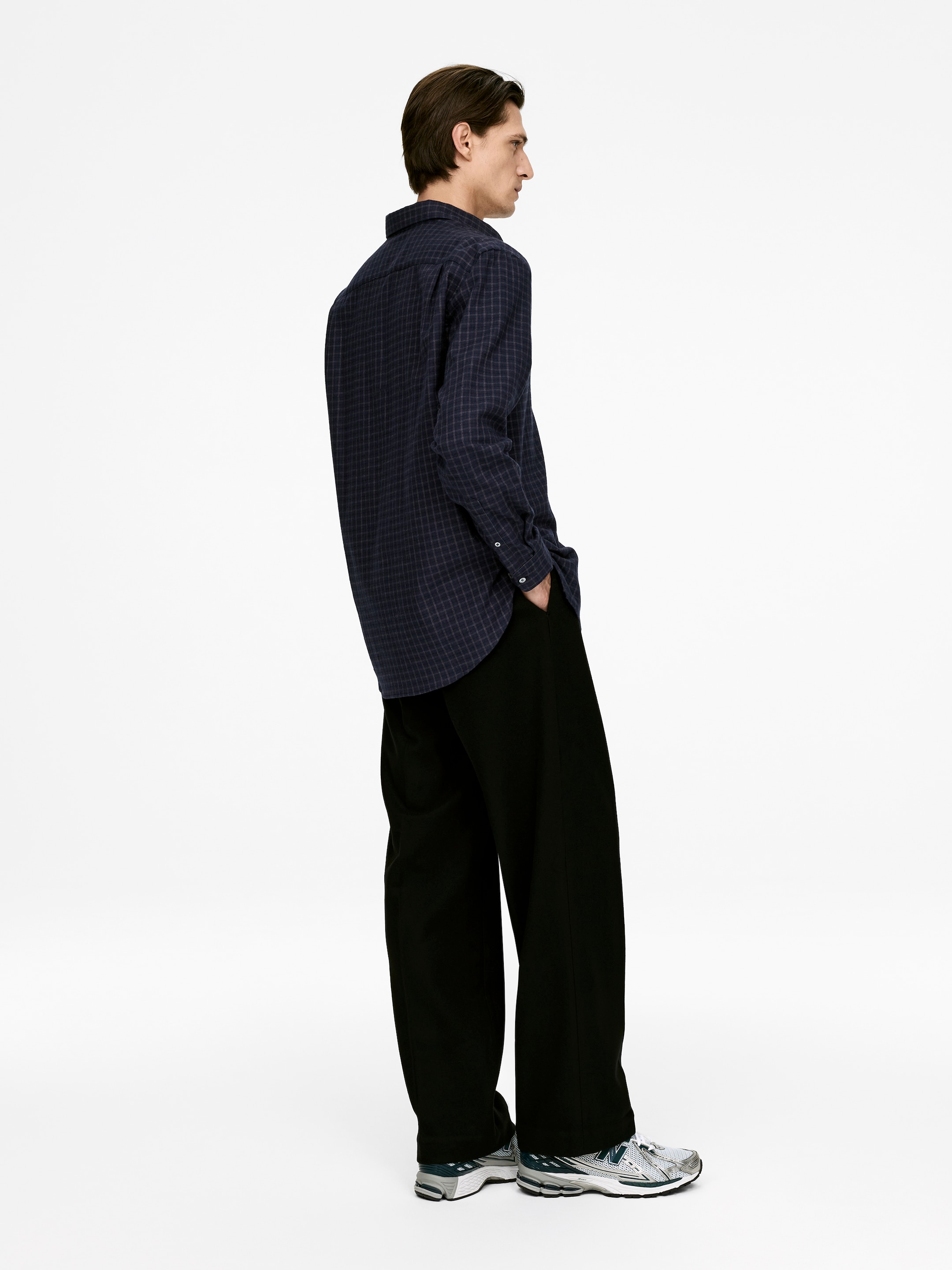 Visualizza immagine più grande: Una camicia blu scuro a maniche lunghe, con un discreto motivo a griglia, viene presentata di lato e di schiena, mettendo in evidenza il colletto classico, i polsini con bottoni e l'orlo che cade liberamente su pantaloni ampi neri.