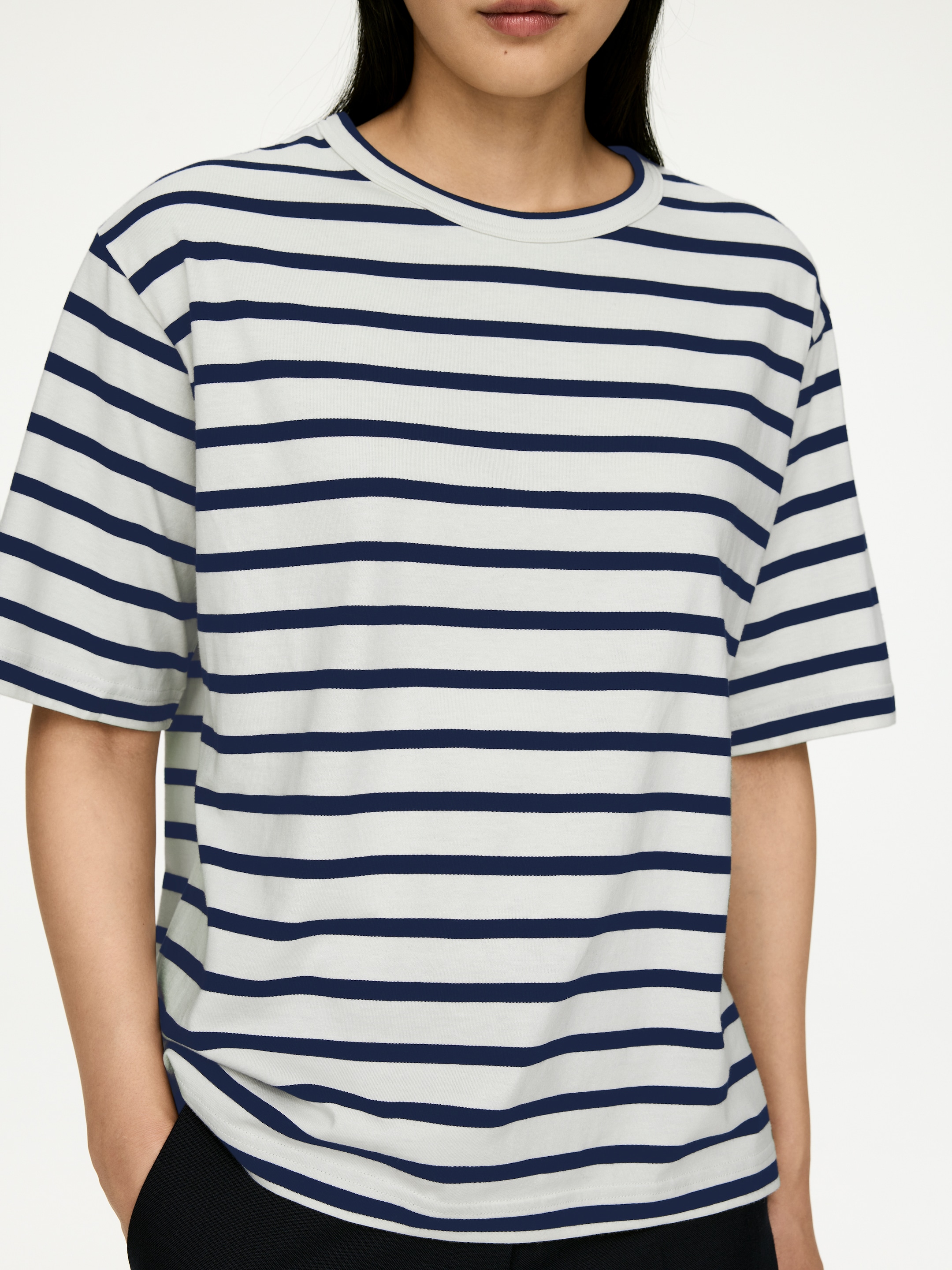 Vis større bilde: T-skjorte i middels vekt i hvit med horisontale marineblå striper, med rund hals og korte ermer. T-skjorten bæres av modellen, med en avslappet passform som henger over svarte bukser mens en av modellens hender hviler i en lomme.